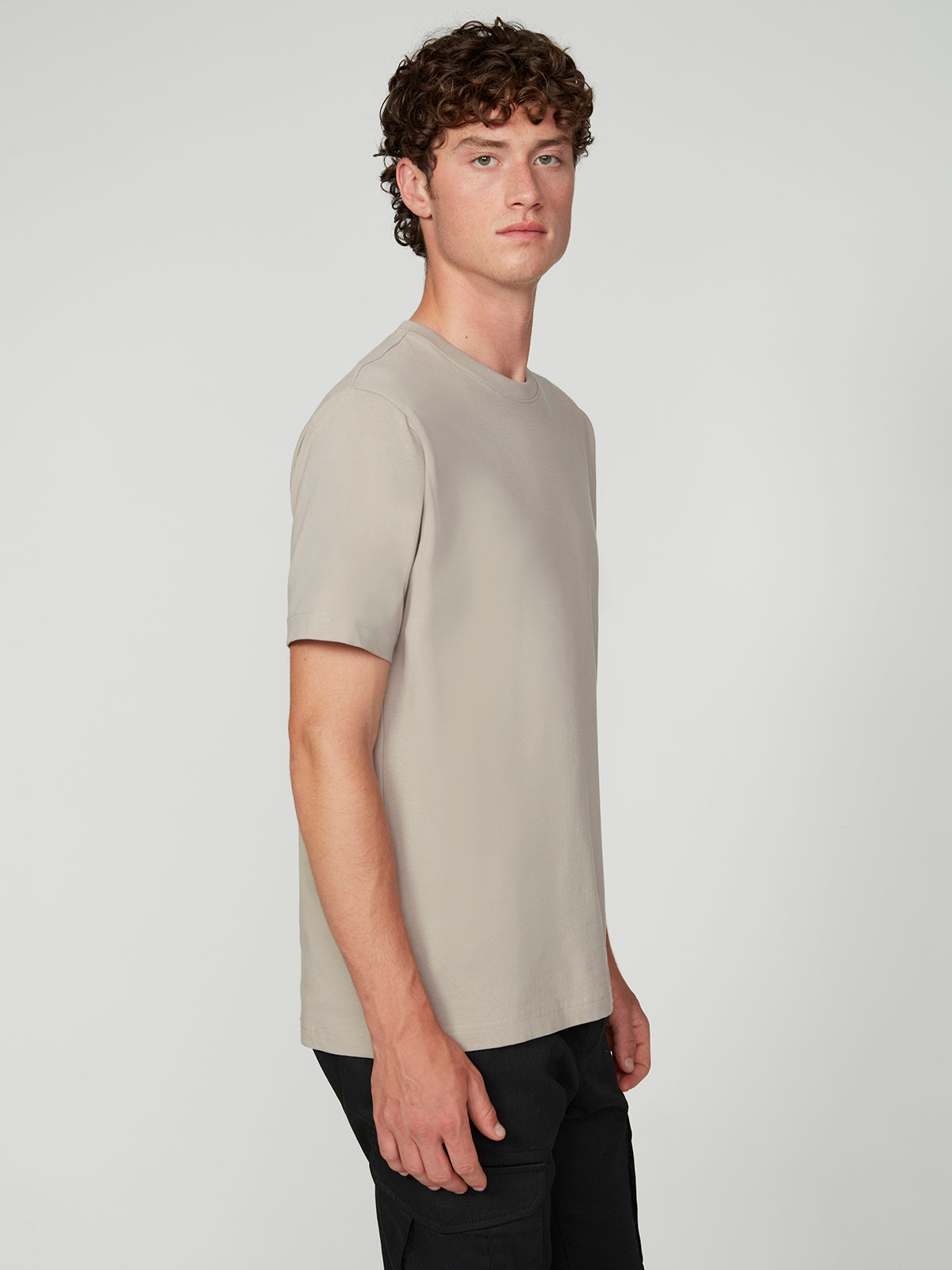 A young man with curly brown hair stands sideways against a light gray background, wearing the PROJEK RAW MENS SHORT SLEEVE CREW NECK T-SHIRT in beige and black pants, looking toward the camera with a neutral expression—perfect for casual style.