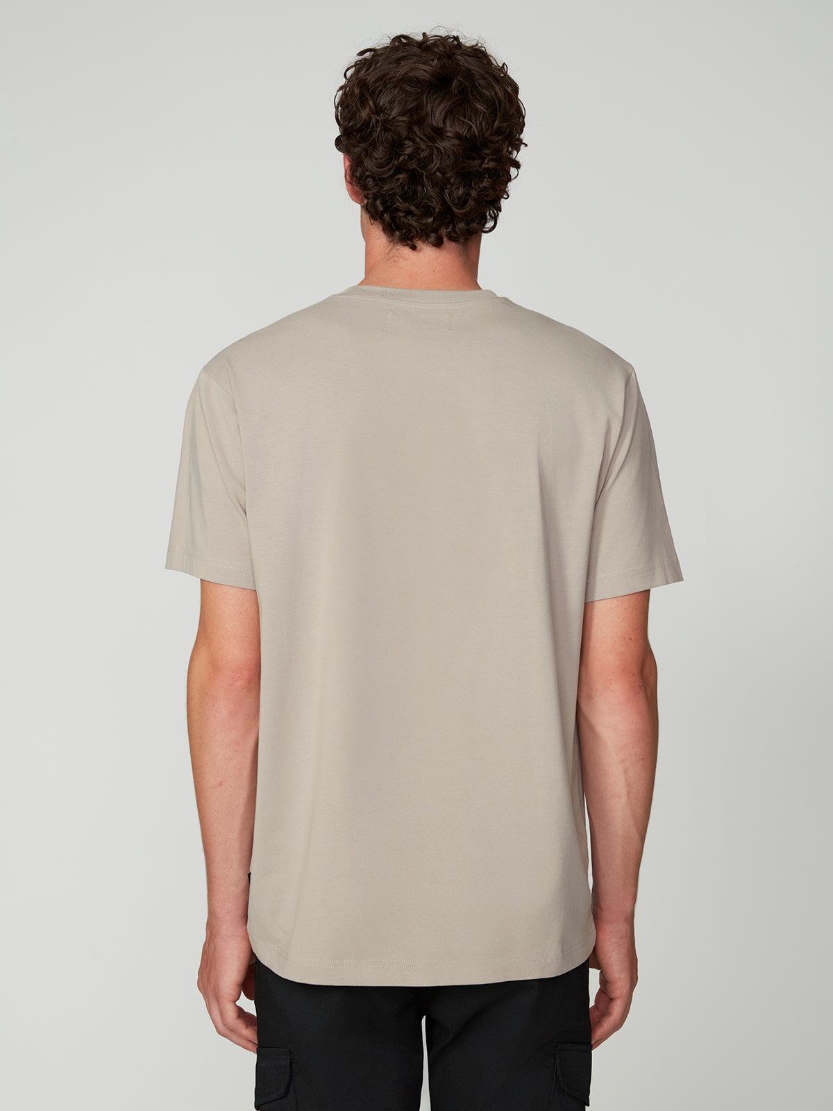 A person with short curly hair faces away, wearing a PROJEK RAW MENS SHORT SLEEVE CREW NECK T-SHIRT in light beige and black pants, standing against a plain light gray background.