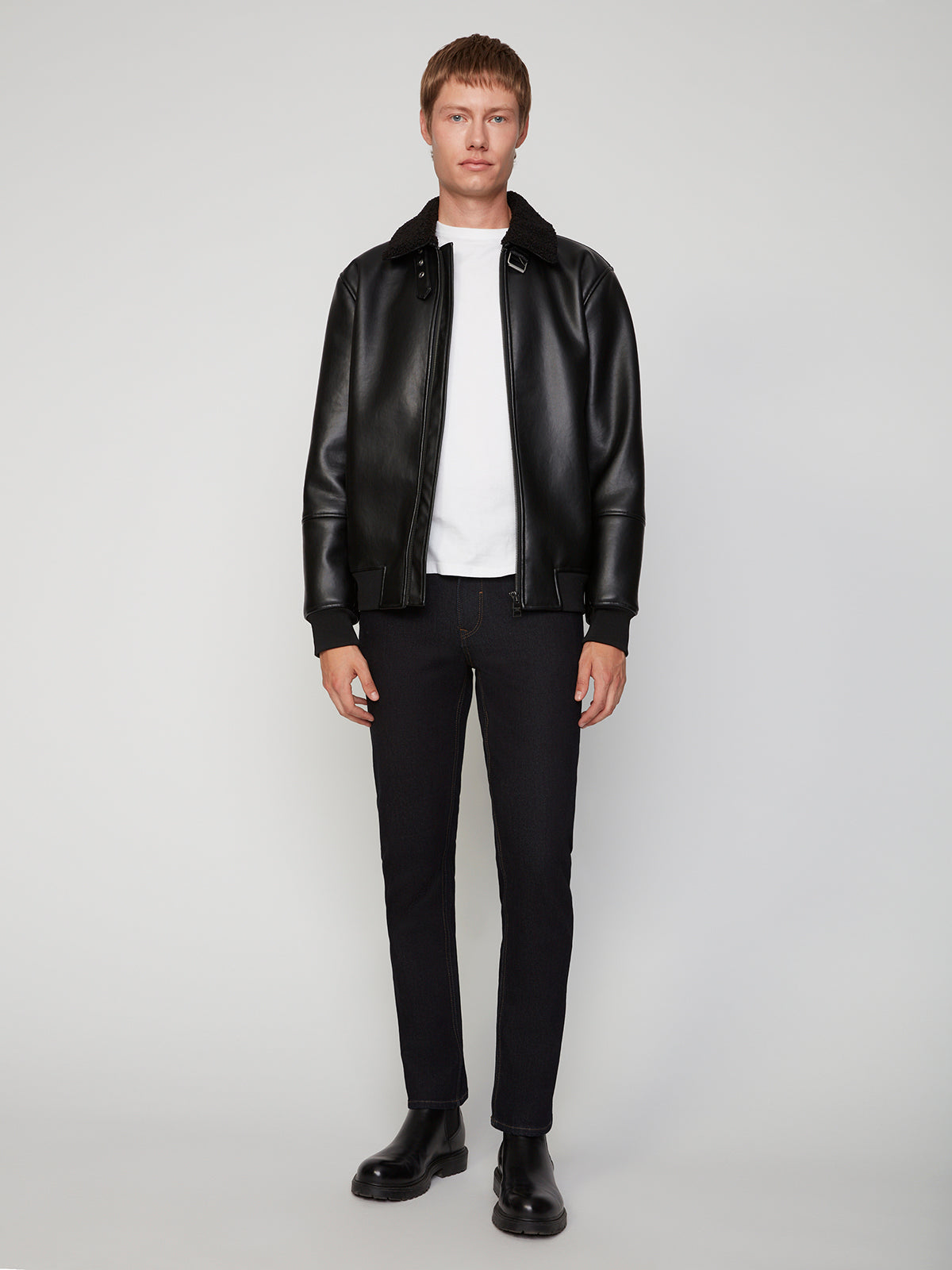 A person stands against a plain background, wearing a black leather jacket, white t-shirt, PROJEK RAW MENS STRETCH SLIM FIT JEAN in black, and black boots, facing forward with a neutral expression.