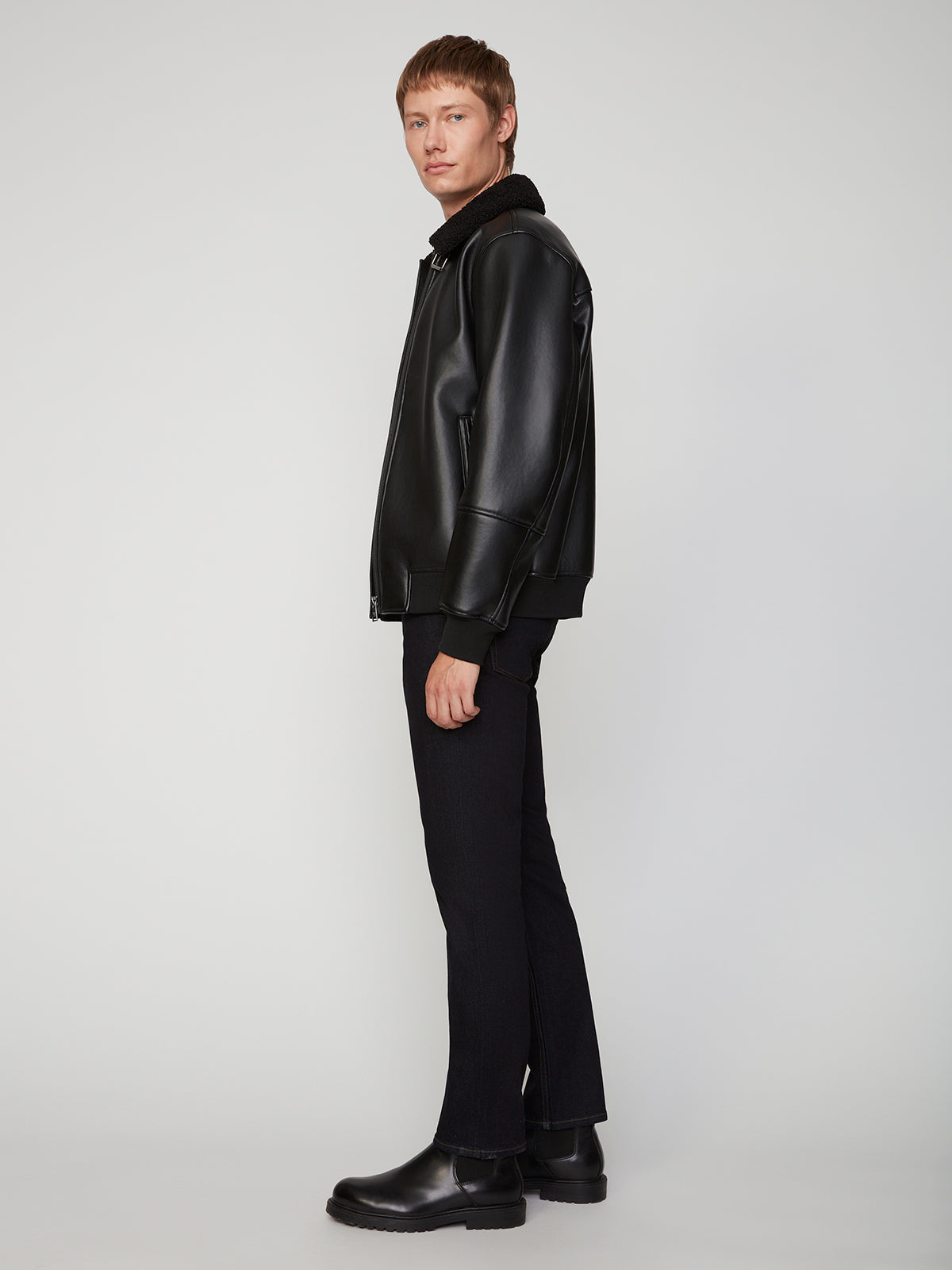 A person with short light brown hair stands sideways, wearing a black leather jacket, PROJEK RAW MENS STRETCH SLIM FIT JEAN, and black boots against a plain light gray background.