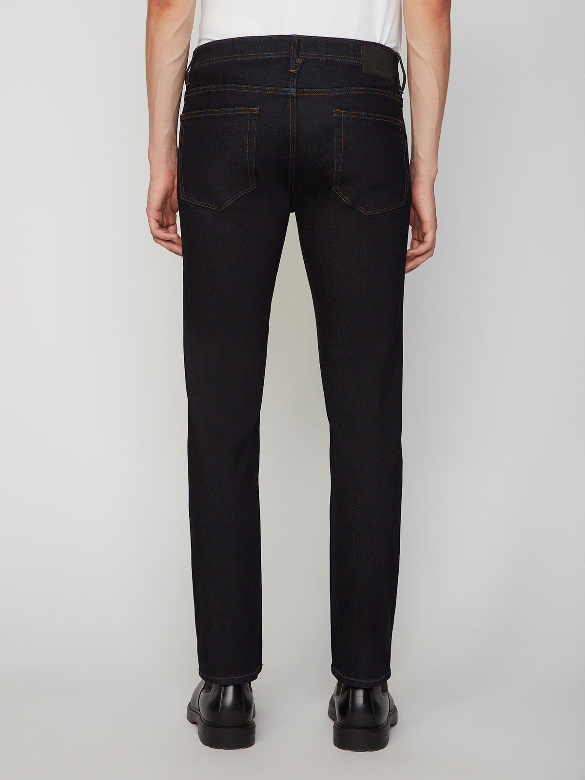 A person in PROJEK RAW MENS STRETCH SLIM FIT JEAN, paired with a white shirt and black shoes, stands facing away from the camera, showcasing the modern slim silhouette against a plain light background.