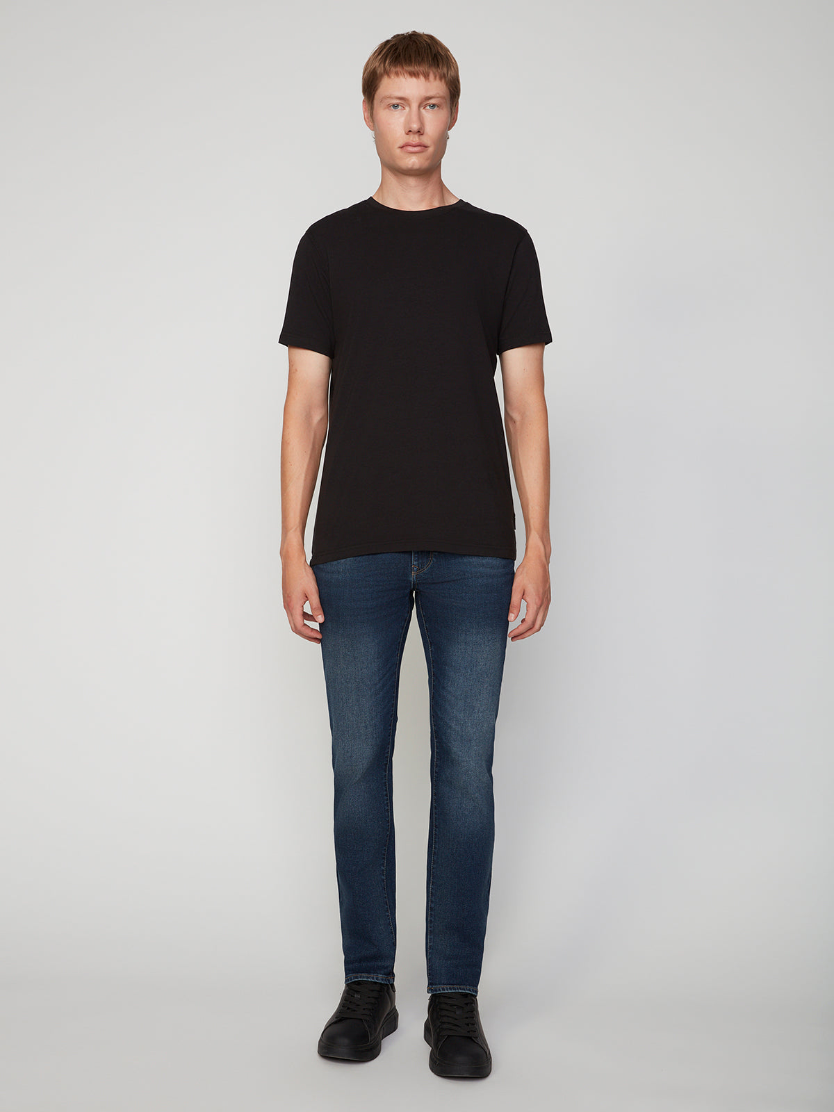 A person stands facing forward against a plain light background, wearing a black short-sleeve t-shirt, PROJEK RAW MENS BARU STRAIGHT FIT JEAN in stretch slub denim, and black shoes.