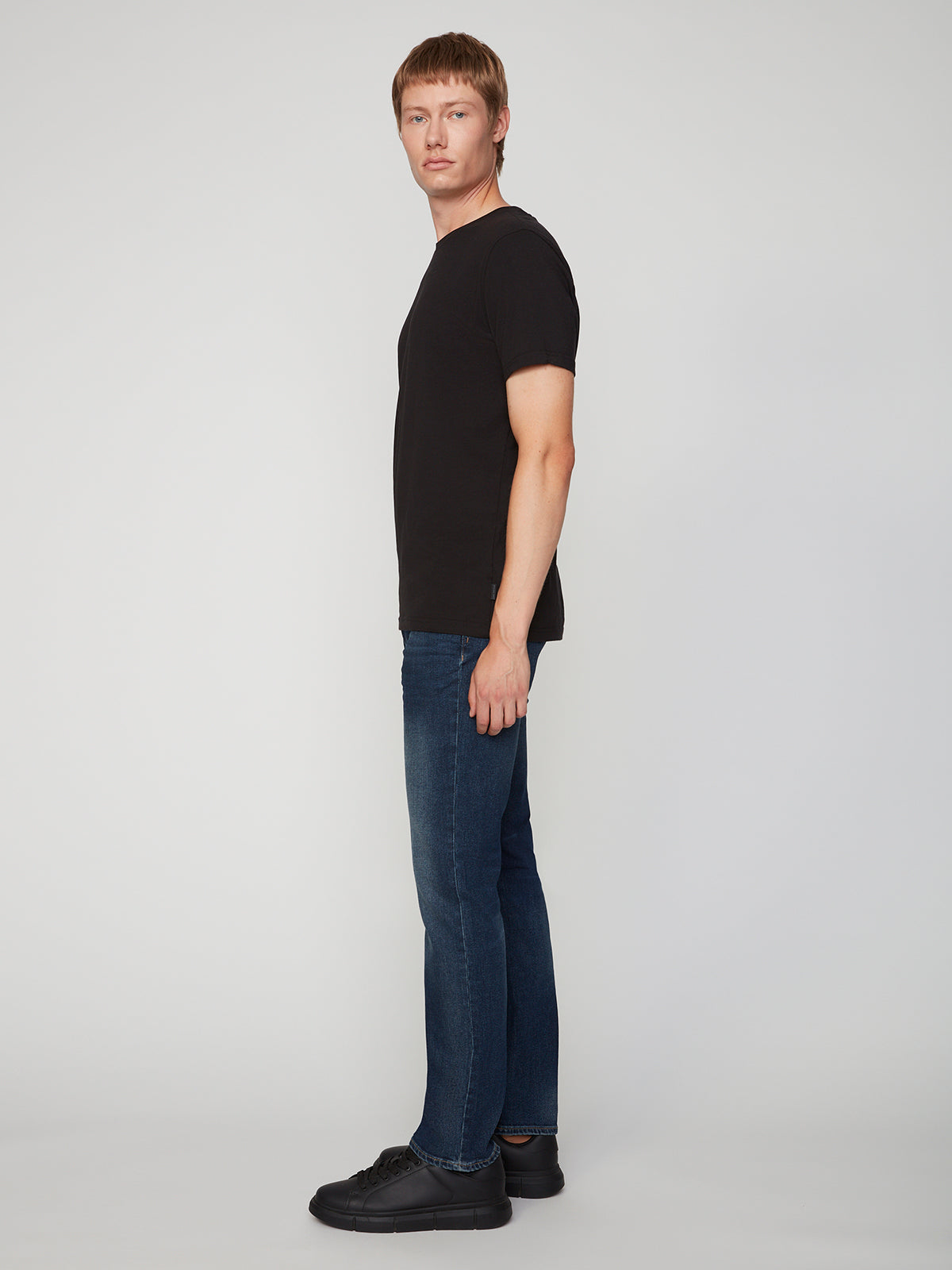 A person with short hair stands sideways against a light gray background, wearing a black t-shirt, black sneakers, and PROJEK RAW MENS BARU STRAIGHT FIT JEAN.