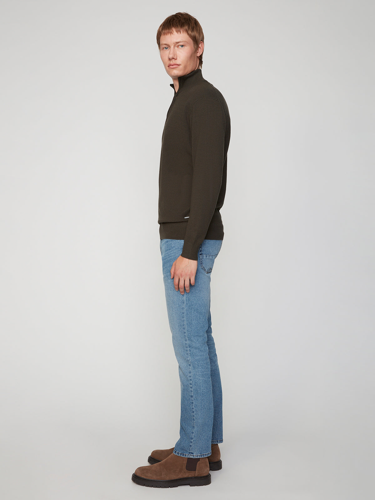 A man stands in profile against a plain light background, wearing a dark zip-up sweater, PROJEK RAW MENS BARU STRAIGHT FIT STRETCH JEAN, and brown boots.