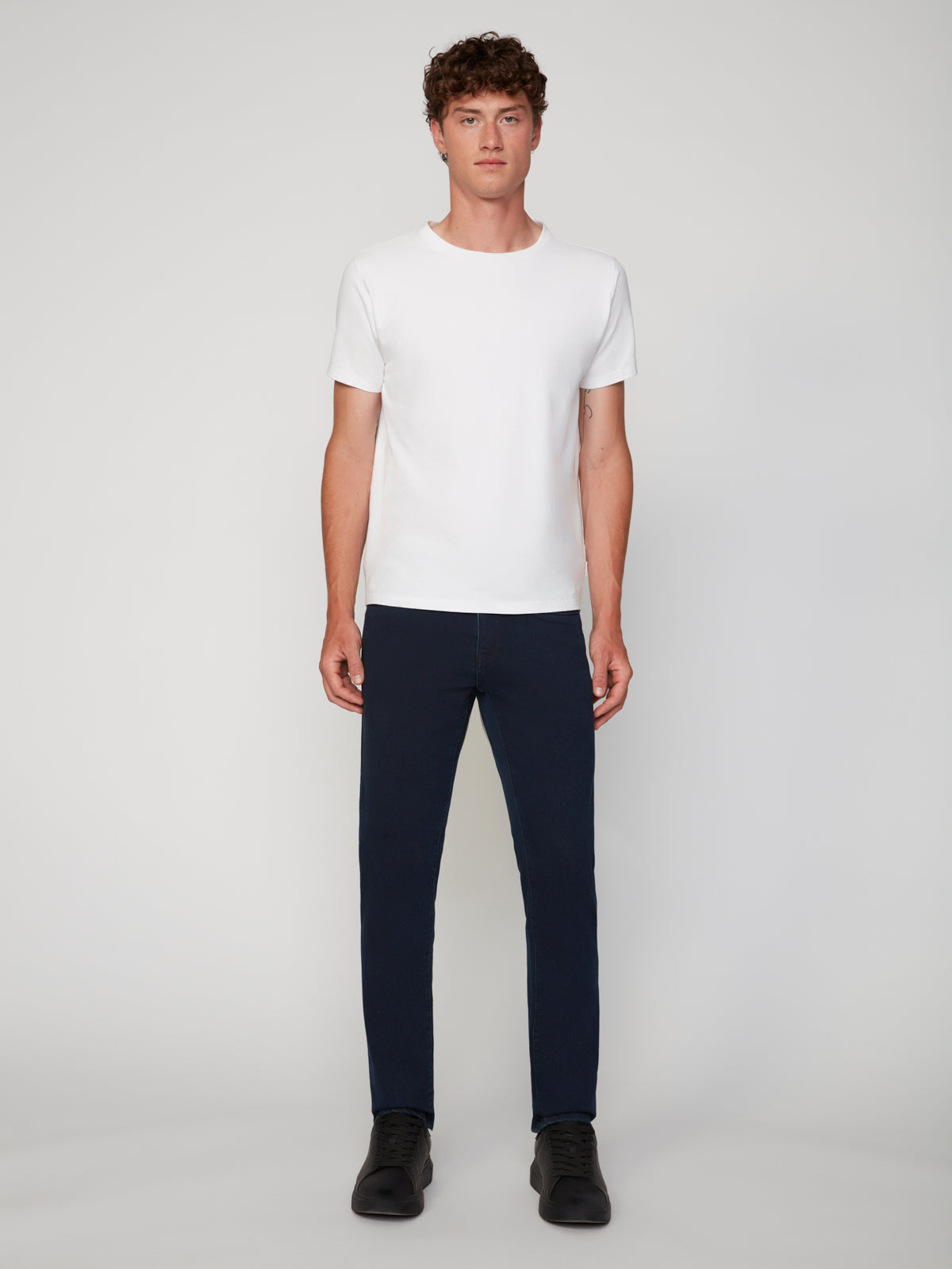 A man with curly hair stands facing forward against a light grey background, wearing a plain white short-sleeve t-shirt, PROJEK RAW MENS BARU STRAIGHT FIT COLORED DENIM JEAN in dark blue, and black shoes.