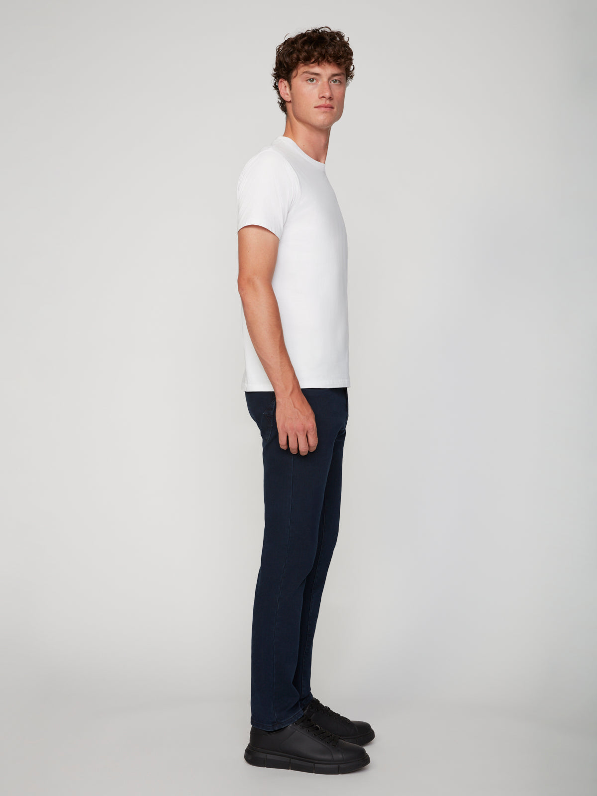 A young man with curly hair stands in profile against a plain light background, wearing a fitted white t-shirt, black sneakers, and PROJEK RAW MENS BARU STRAIGHT FIT COLORED DENIM JEANS.
