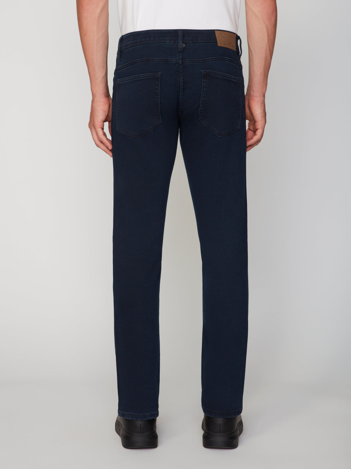 A man wearing PROJEK RAW MENS BARU STRAIGHT FIT COLORED DENIM JEAN in dark blue and black shoes stands with his back to the camera, showing two back pockets and a brown waistband patch against a plain light background.