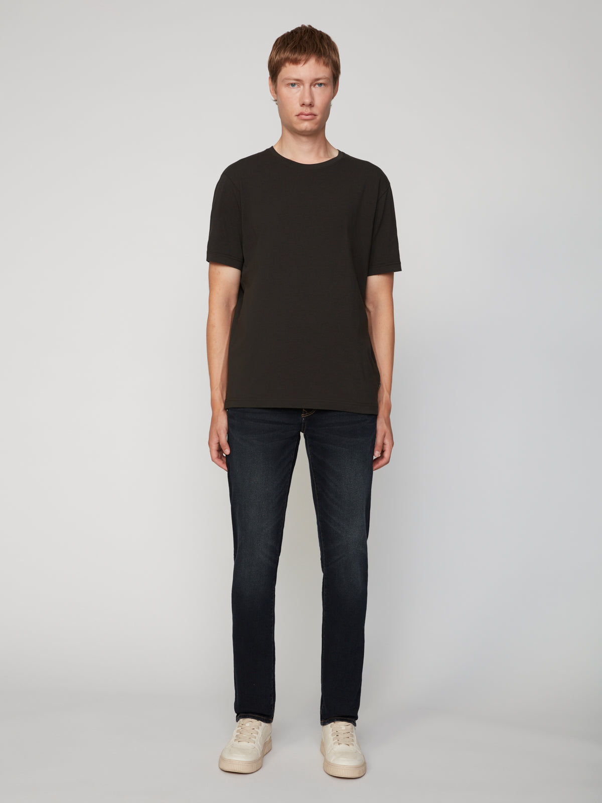 A person with short light brown hair faces forward, wearing a plain black T-shirt, PROJEK RAW MEN’S STRETCH SLUB DENIM BARU STRAIGHT FIT PANT in dark blue, and white sneakers against a plain light gray background.