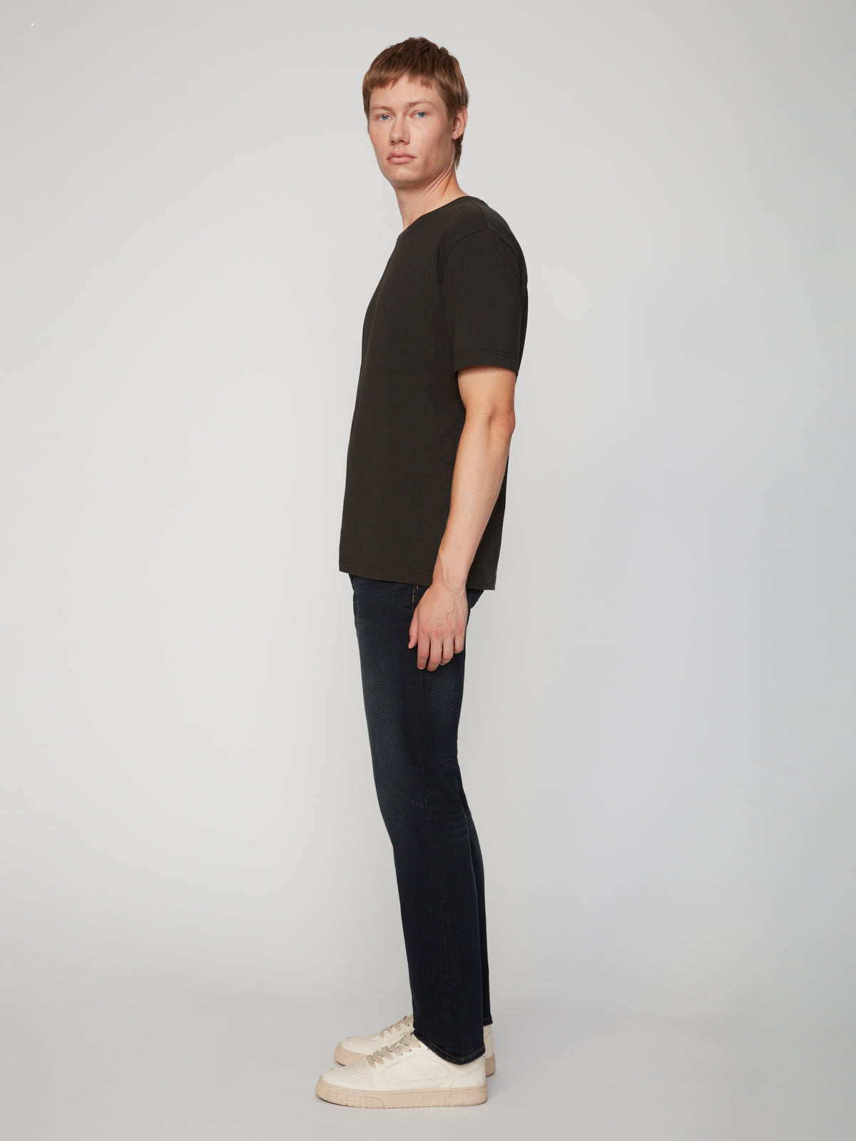 A person with short hair stands in profile against a plain background, wearing a black t-shirt, white sneakers, and PROJEK RAW MEN’S STRETCH SLUB DENIM BARU STRAIGHT FIT PANT.