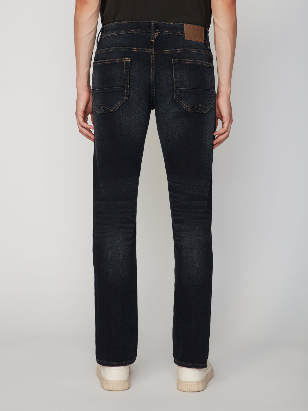 A person wearing PROJEK RAW MEN’S STRETCH SLUB DENIM BARU STRAIGHT FIT PANT in dark blue and white shoes stands facing away, highlighting the back pockets and a brown waistband patch.