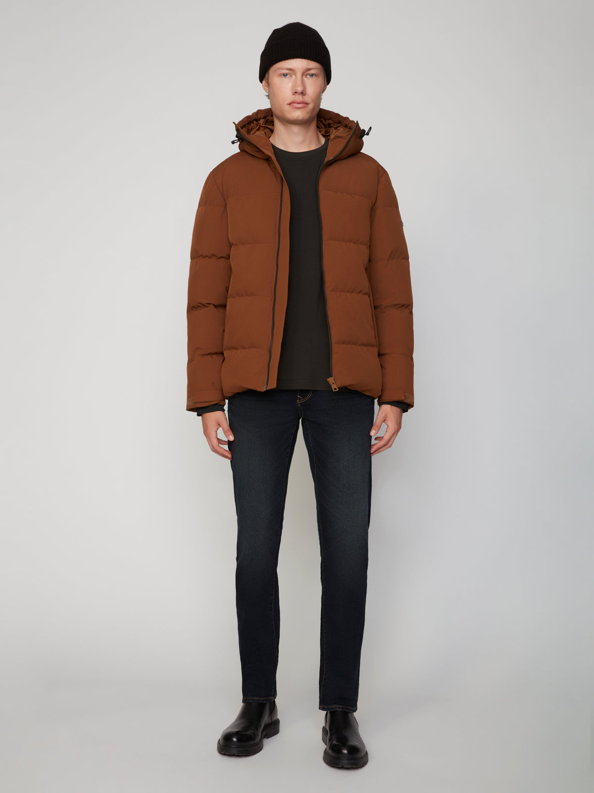 A man faces forward in a PROJEK RAW MENS PUFFER JACKET WITH HOOD, paired with a black beanie, dark shirt, slim jeans, and black boots against a plain light background—highlighting lightweight warmth and modern style.