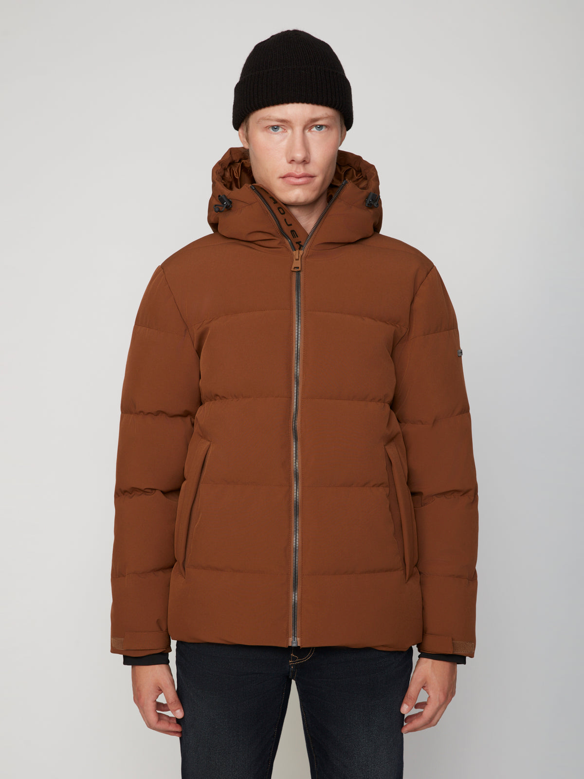 A man in the PROJEK RAW MENS PUFFER JACKET WITH HOOD and a black beanie stands before a light gray backdrop, showcasing lightweight warmth ideal for chilly days.