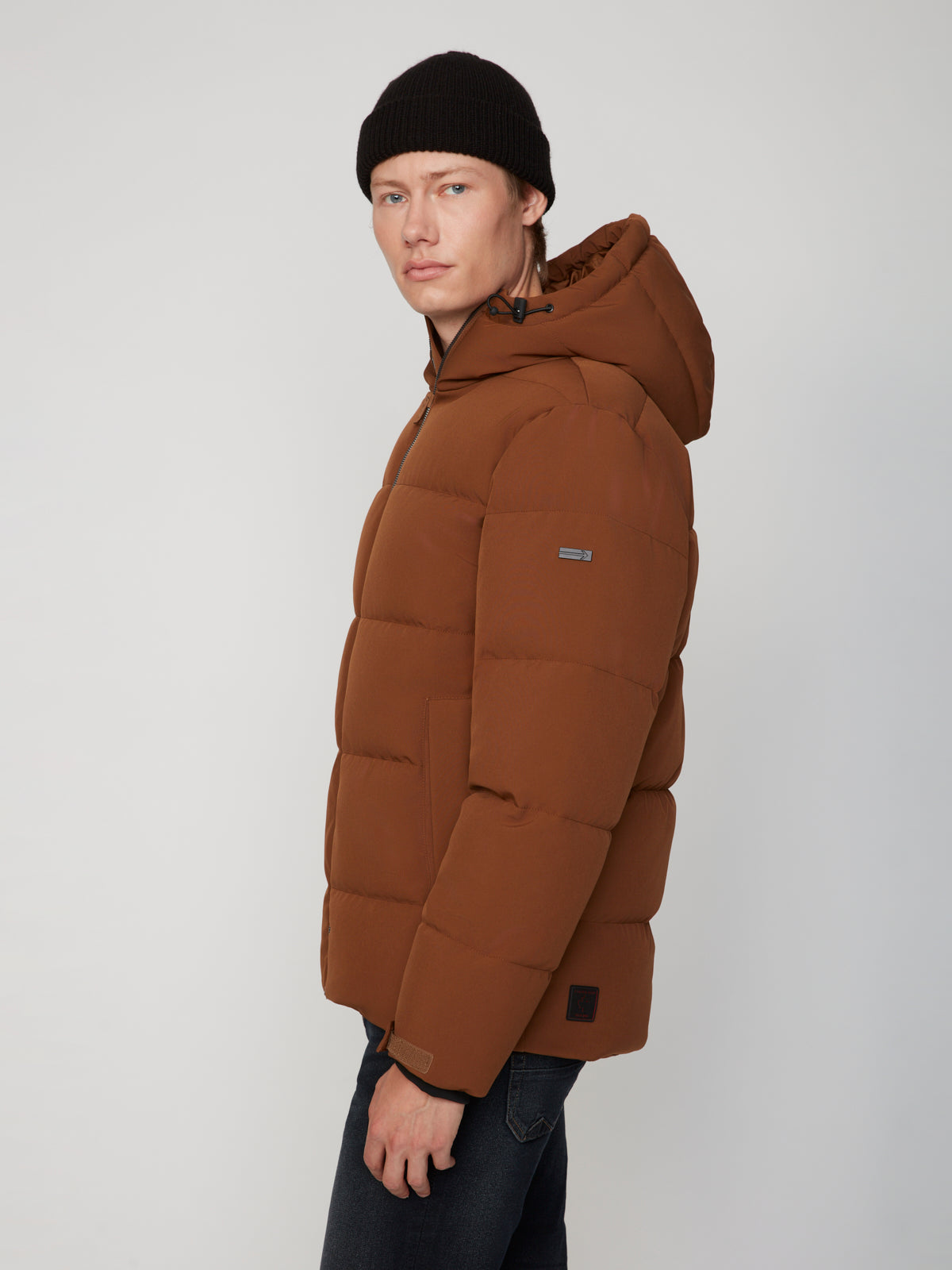 A man wears the PROJEK RAW MENS PUFFER JACKET WITH HOOD in brown and a black knit beanie, standing against a plain light background—ideal for lightweight warmth on chilly days.