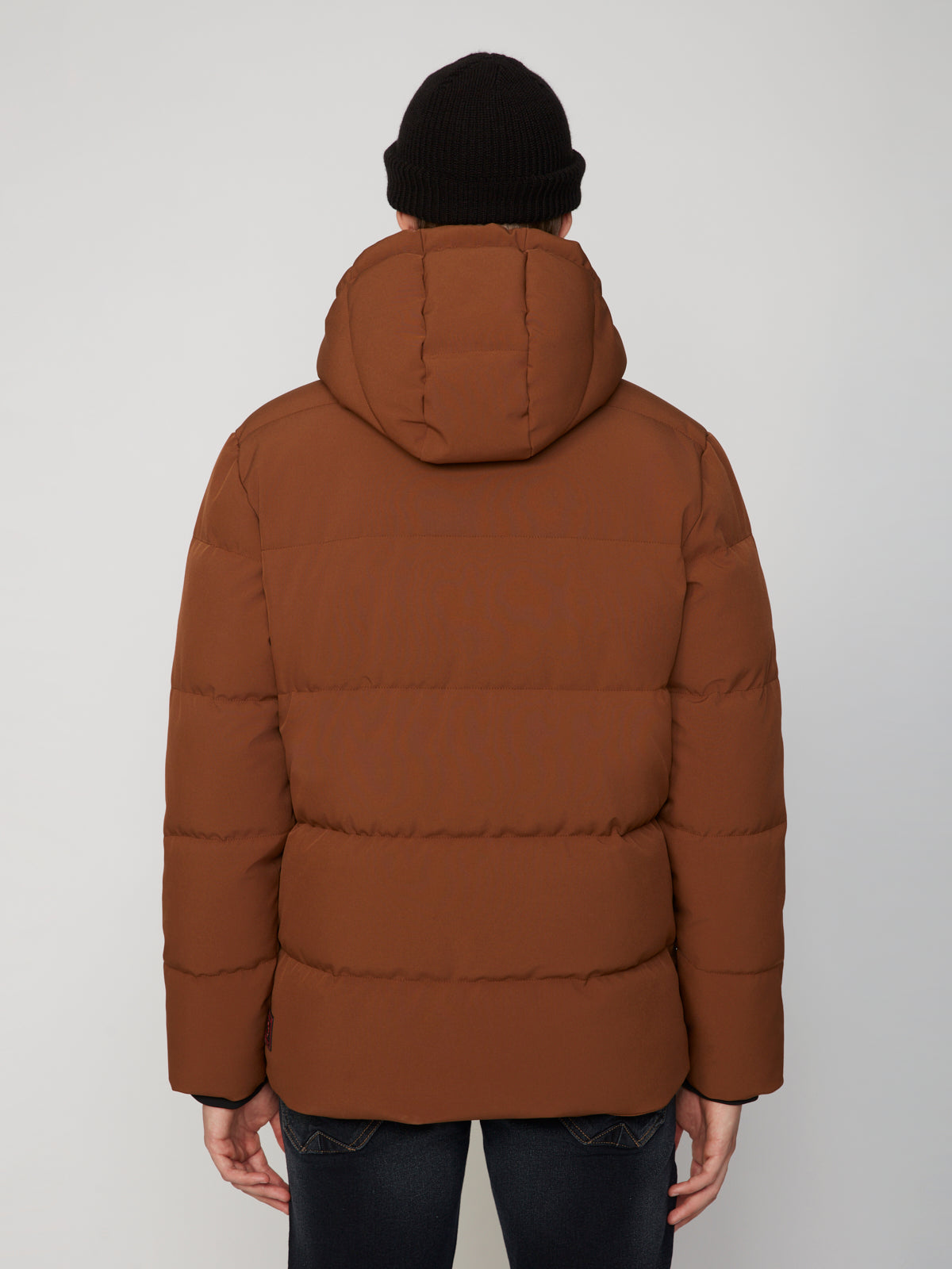 A person facing away wears the PROJEK RAW MENS PUFFER JACKET WITH HOOD in brown and a black knit beanie, set against a plain, light background to highlight its lightweight warmth.