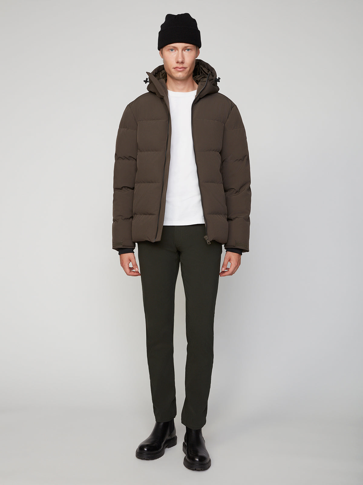 A person wearing a black beanie, PROJEK RAW MENS PUFFER JACKET WITH HOOD in brown, white t-shirt, dark green pants, and black boots stands against a plain light gray background.