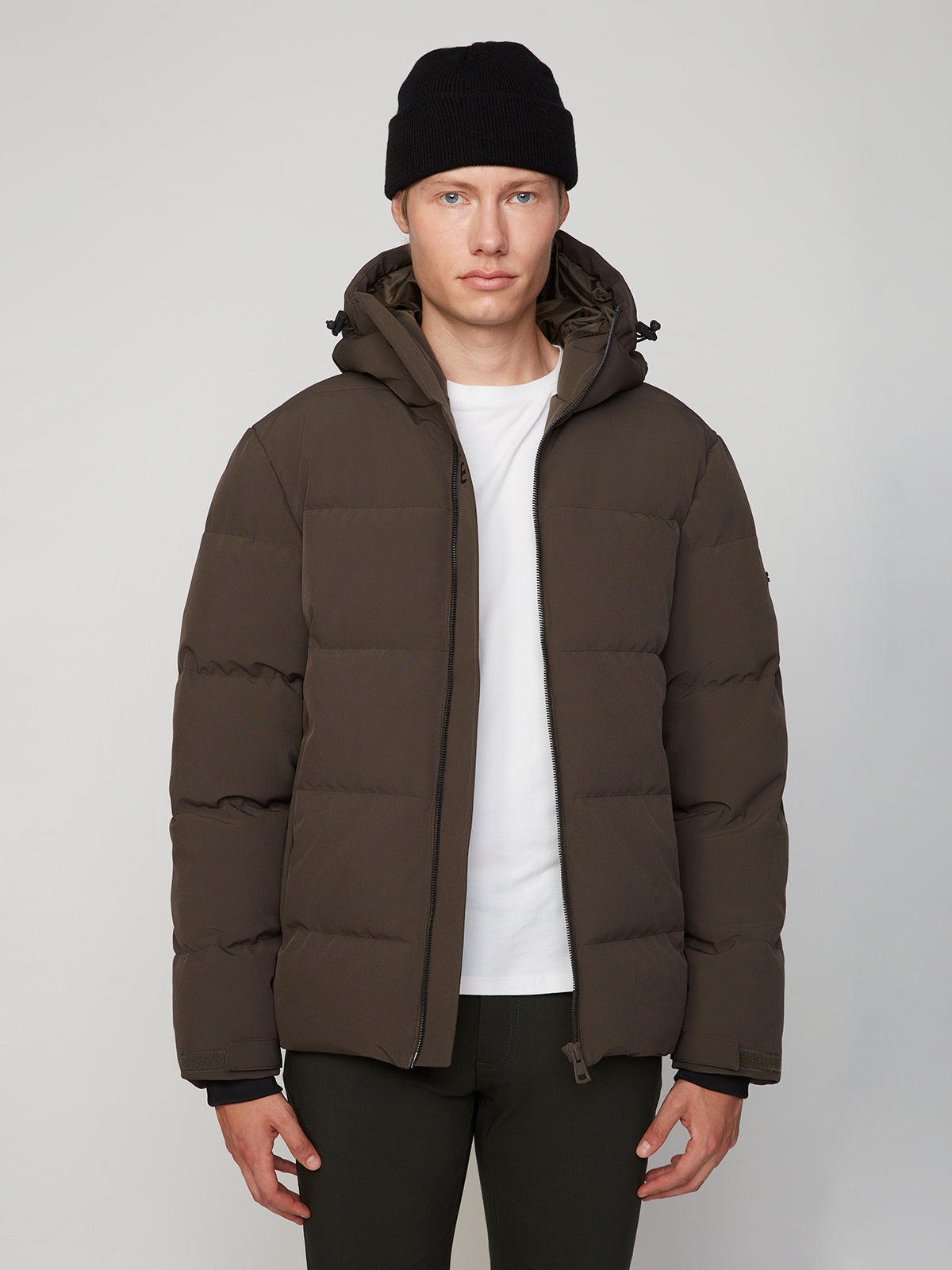 A person wearing a black beanie, white t-shirt, dark pants, and the PROJEK RAW MENS PUFFER JACKET WITH HOOD in dark brown stands against a plain light background, looking directly at the camera.