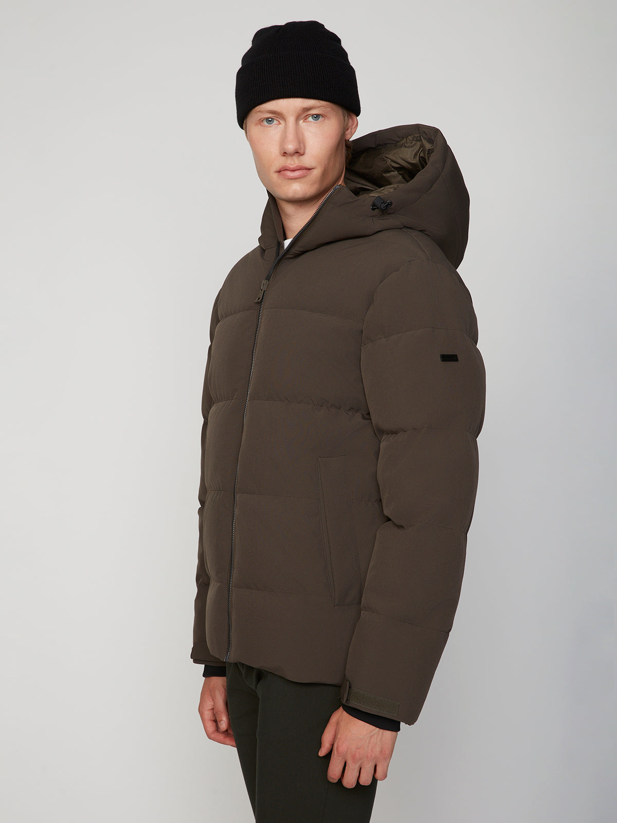 A young man in a black beanie and the PROJEK RAW MENS PUFFER JACKET WITH HOOD stands against a light gray background, looking at the camera. This insulated jacket delivers lightweight warmth for chilly days.
