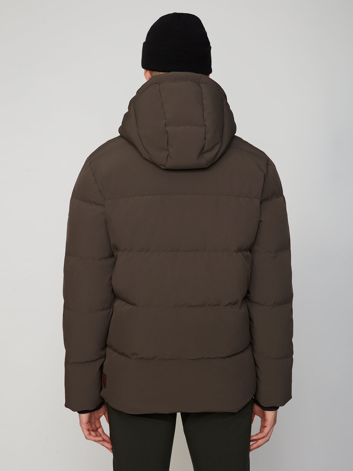 A man wears the PROJEK RAW MENS PUFFER JACKET WITH HOOD in dark brown and a black beanie, shown from the back against a light gray background—ideal for lightweight warmth on chilly days.