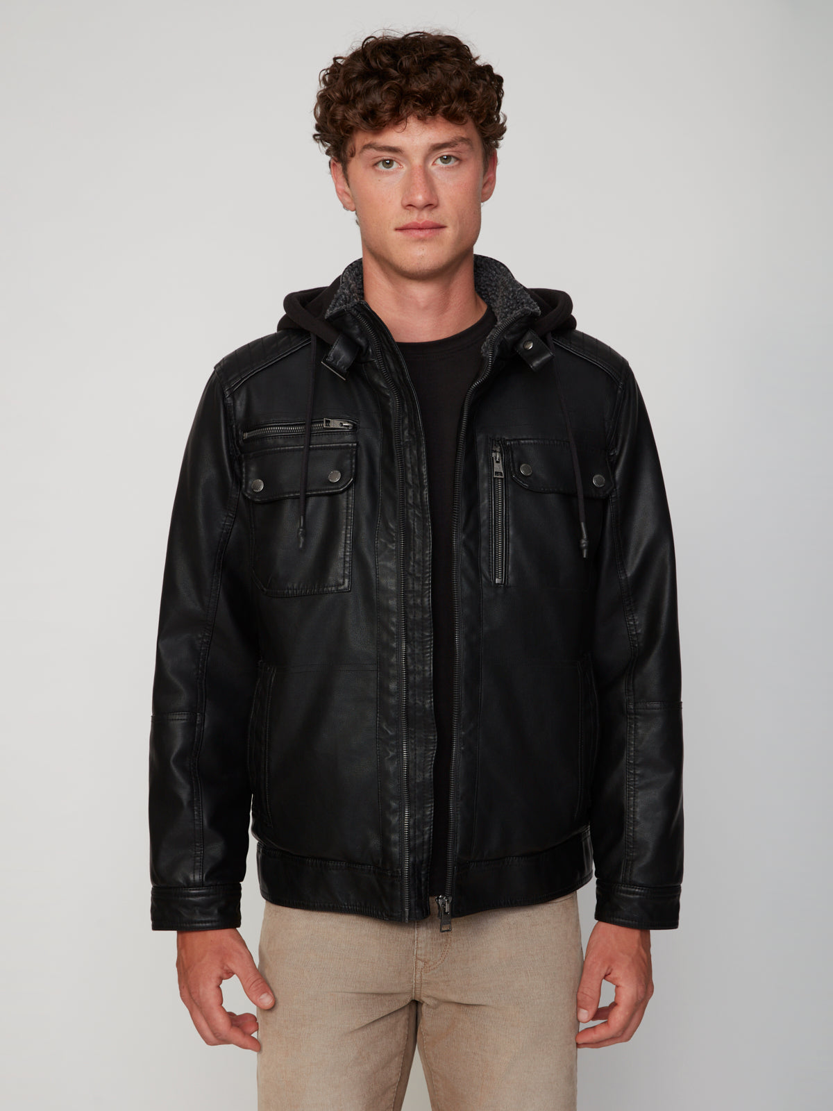 A young man with curly brown hair wears the PROJEK RAW MEN’S VEGAN LEATHER JACKET WITH REMOVABLE HOOD, a black shirt, and beige pants, standing against a light gray background and facing the camera with a neutral expression.