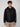 A young man with curly brown hair wears the PROJEK RAW MEN’S VEGAN LEATHER JACKET WITH REMOVABLE HOOD, a black shirt, and beige pants, standing against a light gray background and facing the camera with a neutral expression.