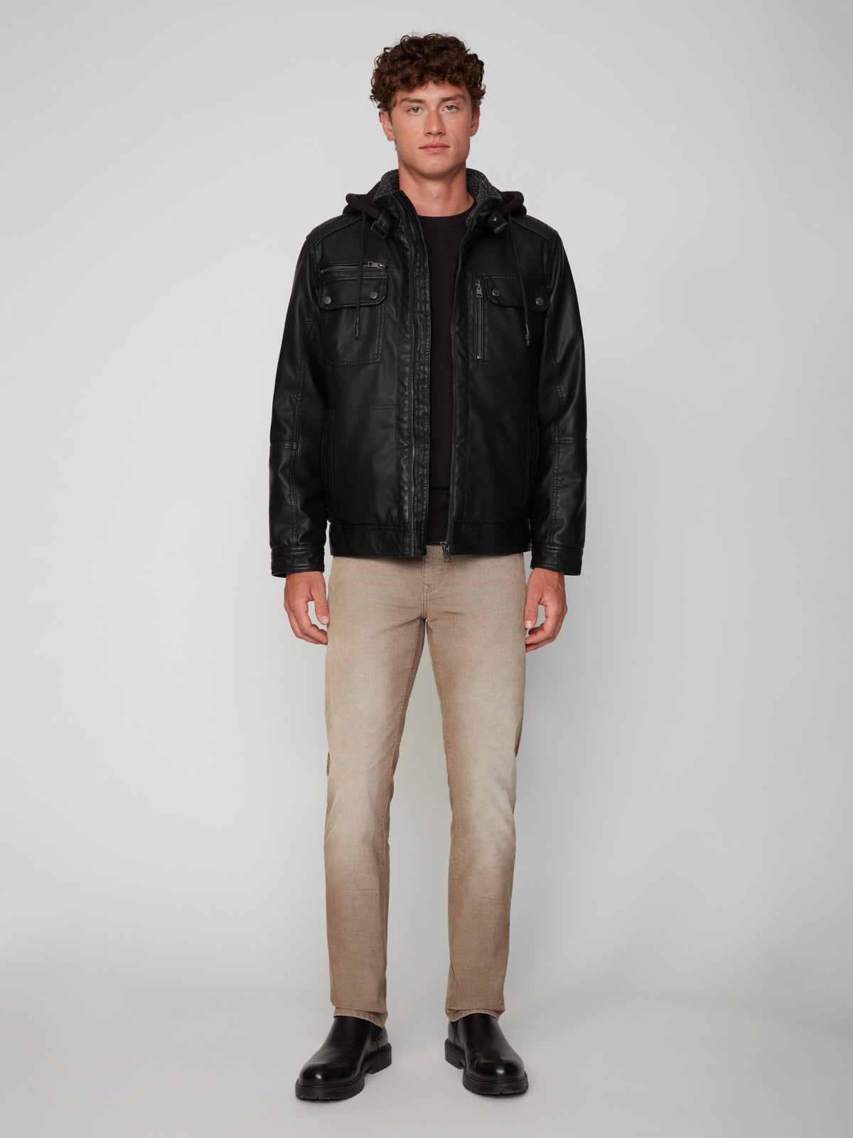 A man faces forward in a PROJEK RAW MEN’S VEGAN LEATHER JACKET WITH REMOVABLE HOOD over a black shirt, light beige pants, and black boots, set against a plain, light background.
