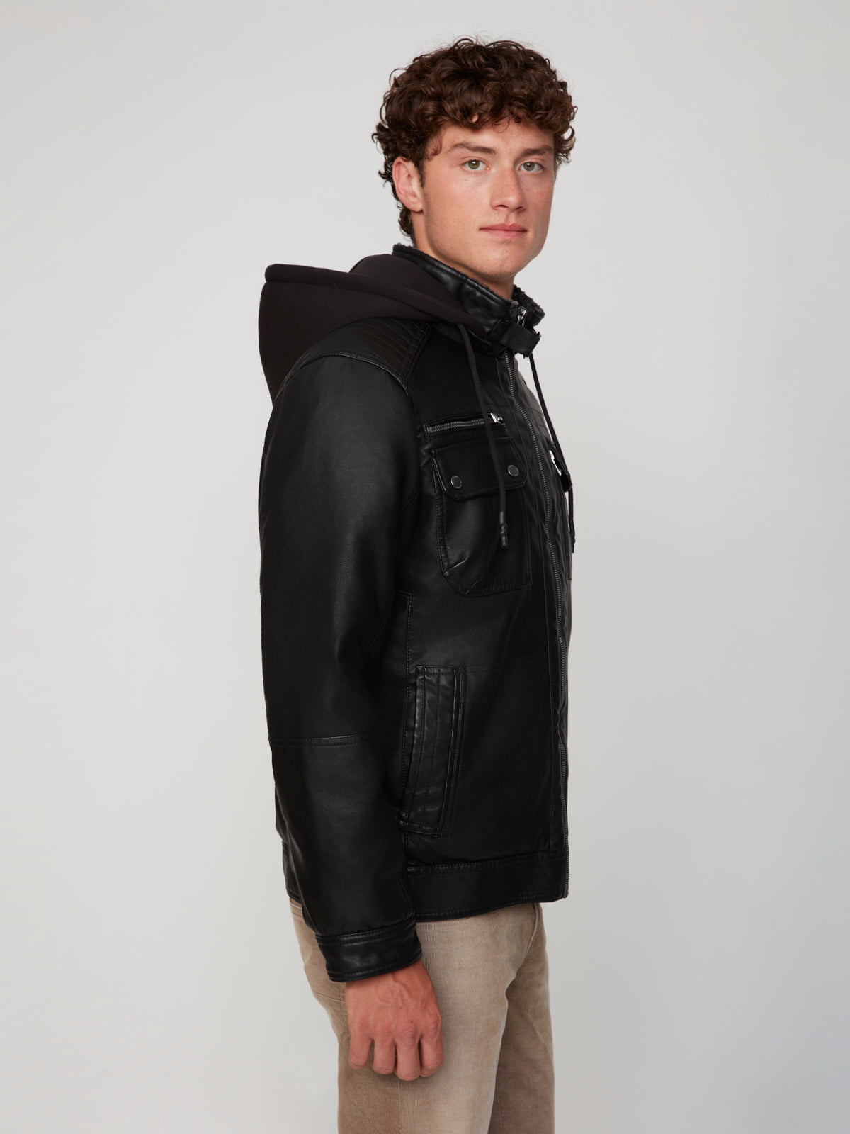 A young man with curly brown hair wears the PROJEK RAW MEN’S VEGAN LEATHER JACKET WITH REMOVABLE HOOD and tan pants, standing sideways and looking at the camera against a plain light background.
