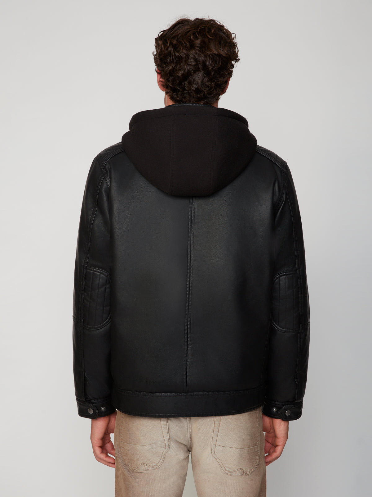 A person with short curly brown hair, seen from behind, wears the PROJEK RAW MEN’S VEGAN LEATHER JACKET WITH REMOVABLE HOOD and light beige pants, standing against a plain light background.