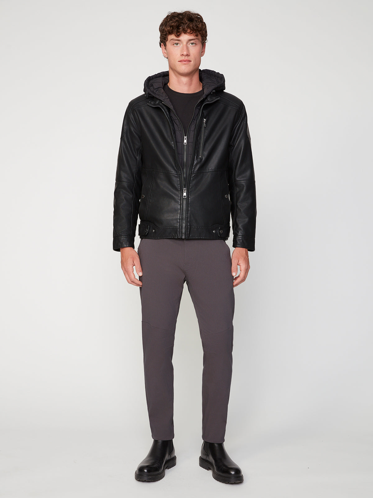 A young man faces forward wearing the PROJEK RAW MENS MOTO JACKET WITH REMOVABLE HOODED DICKIE, styled with dark gray pants, a black t-shirt, and black boots against a plain white background.