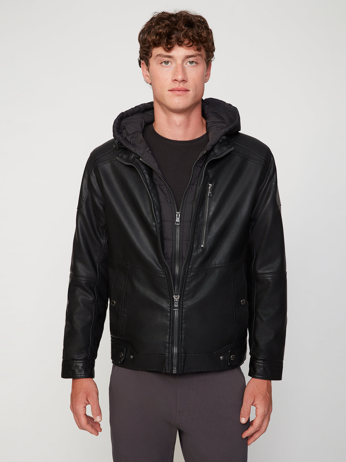 A young man with short curly hair wears the PROJEK RAW MENS MOTO JACKET WITH REMOVABLE HOODED DICKIE over a black shirt and zip-up hoodie, standing facing forward against a plain light gray background with a neutral expression.