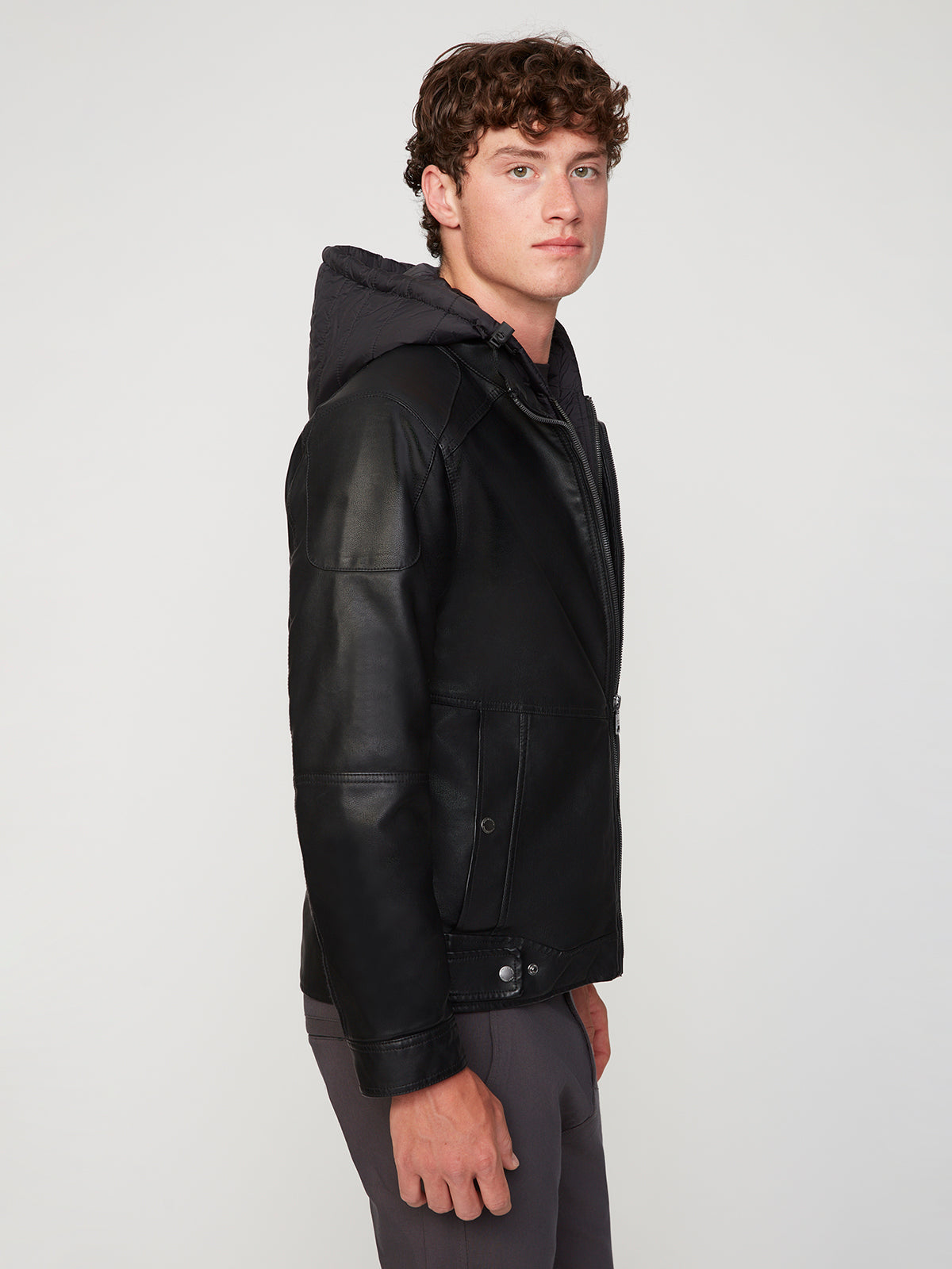 A young man with curly brown hair wears the PROJEK RAW MENS MOTO JACKET WITH REMOVABLE HOODED DICKIE and dark pants, standing sideways and looking at the camera against a plain light background.