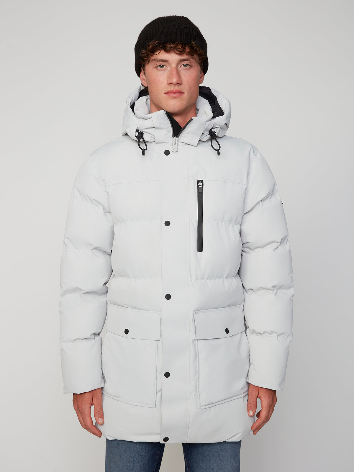 A young man wearing a white PROJEK RAW MENS TEXTURED RIPSTOP ECOFILL QUILTED PARKA WITH HOOD, black beanie, and blue jeans stands facing the camera against a plain light background.