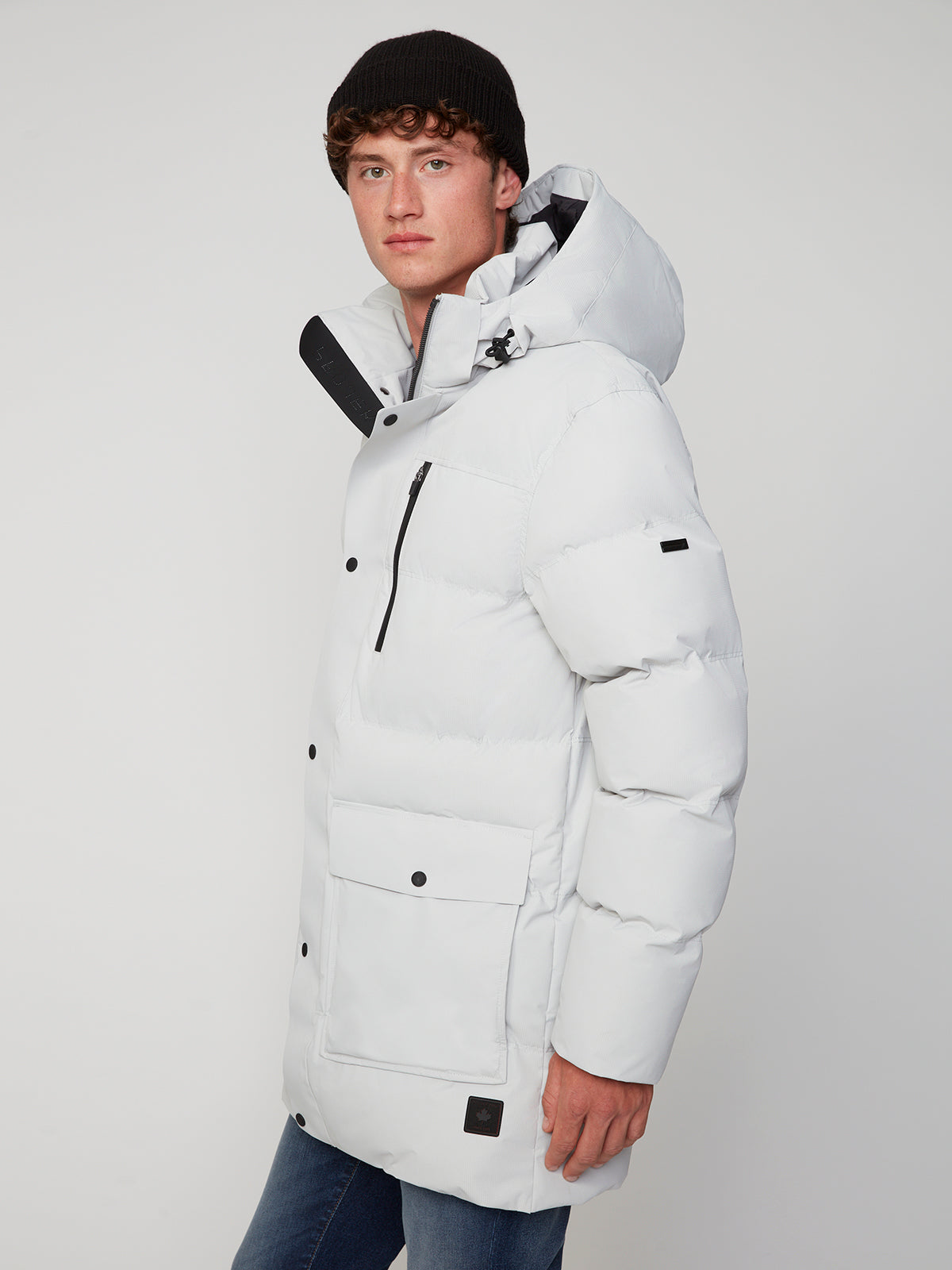 A young man in a black beanie and a white PROJEK RAW MENS TEXTURED RIPSTOP ECOFILL QUILTED PARKA WITH HOOD, featuring multiple pockets and black details, poses against a plain light background, looking slightly to the side.