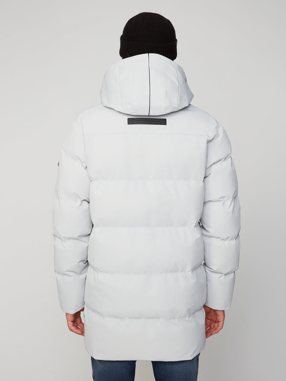 A person is shown from the back wearing a white MENS TEXTURED RIPSTOP ECOFILL QUILTED PARKA WITH HOOD by PROJEK RAW and a black beanie. The mid-thigh-length parka stands out against a plain light gray background.