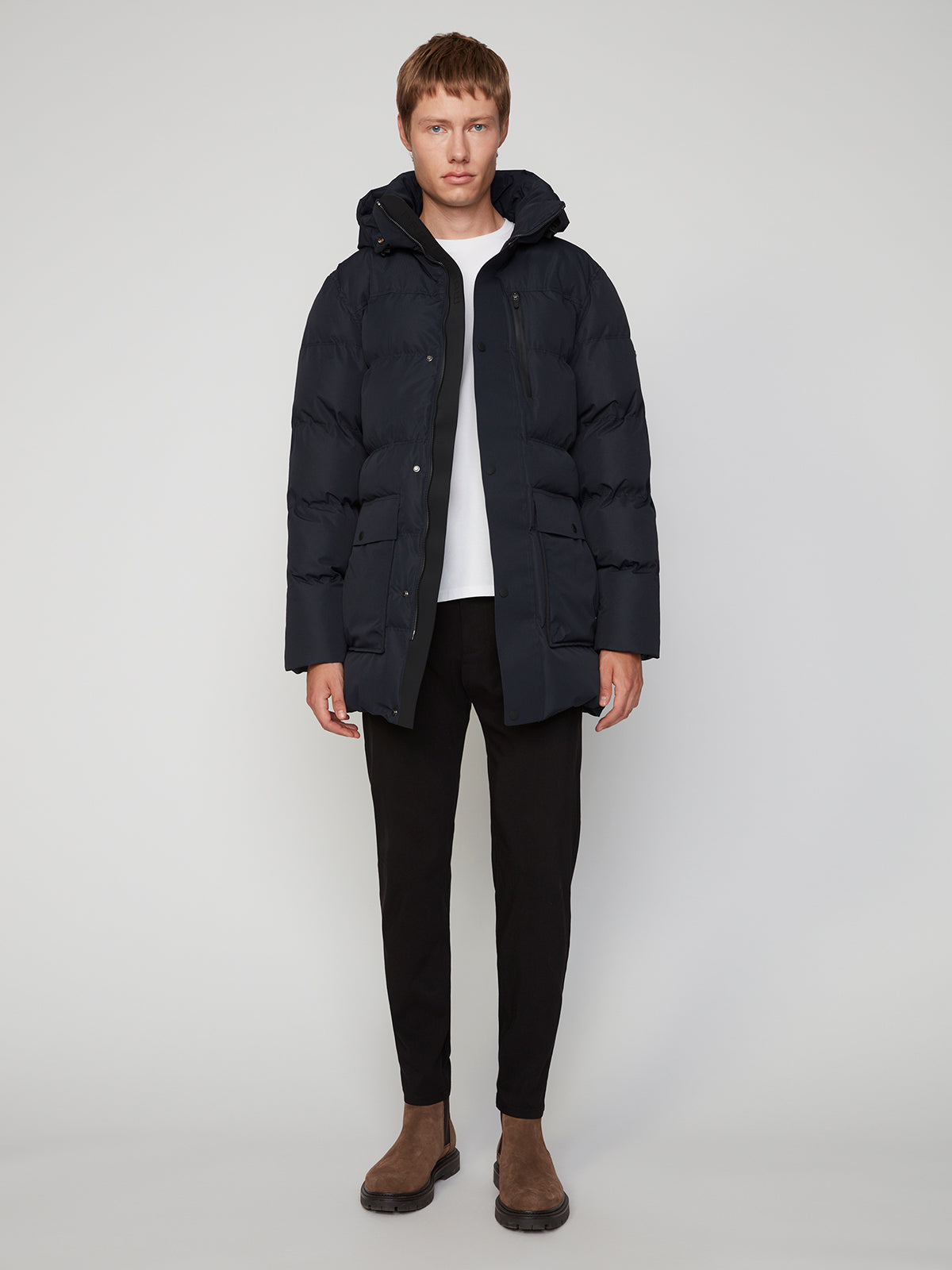 A person faces forward, wearing a PROJEK RAW MENS TEXTURED RIPSTOP ECOFILL QUILTED PARKA WITH HOOD in black, a white shirt, black pants, and brown suede boots against a plain light gray background.
