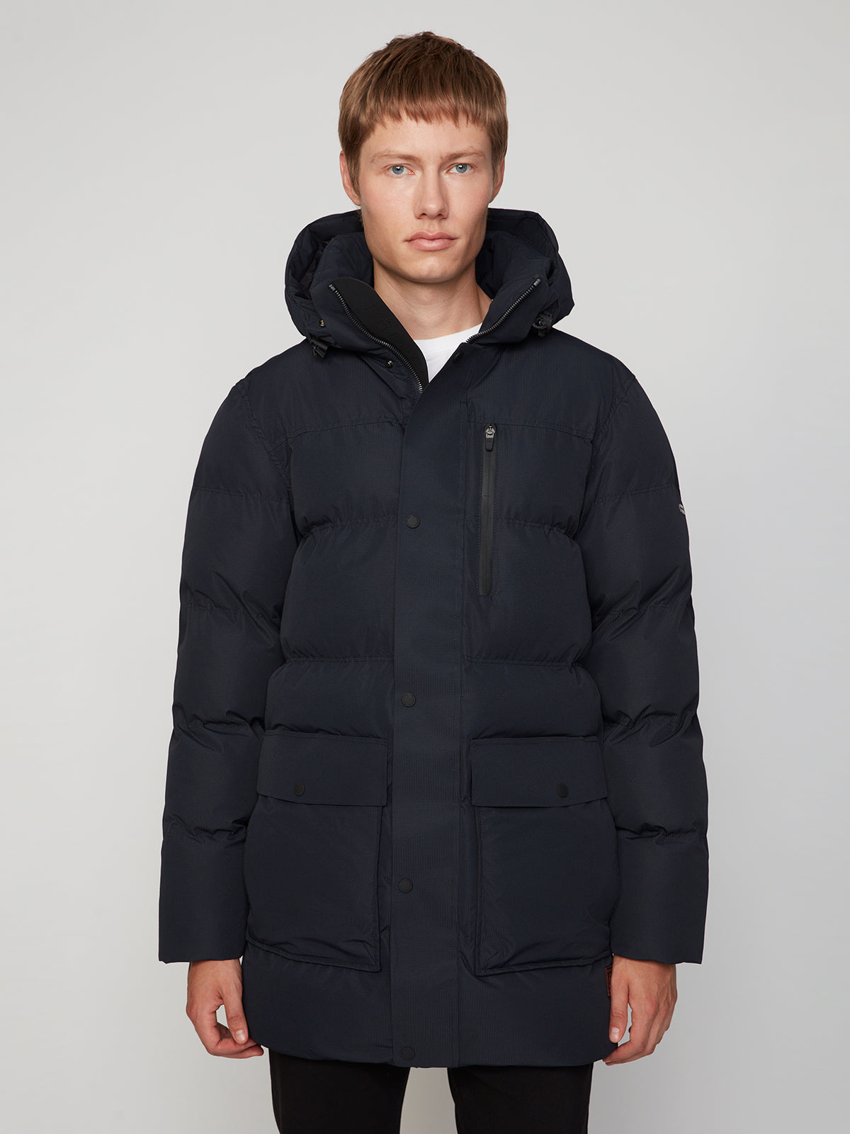 A man with short light brown hair wears the PROJEK RAW MENS TEXTURED RIPSTOP ECOFILL QUILTED PARKA WITH HOOD—an eco-friendly winter coat with large front pockets—facing the camera against a plain light background, expression neutral.