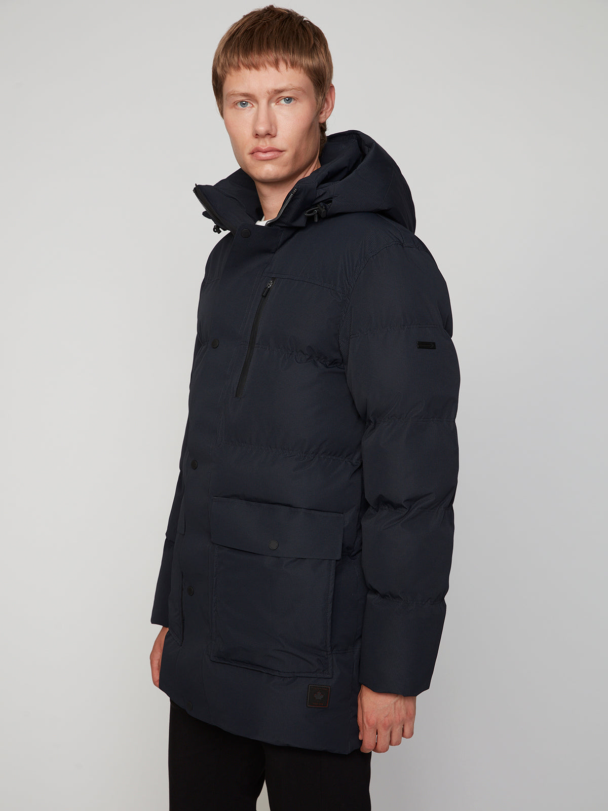 A person with short light brown hair wears the PROJEK RAW MENS TEXTURED RIPSTOP ECOFILL QUILTED PARKA WITH HOOD—a long, dark navy eco-friendly winter coat—standing against a plain light background and looking toward the camera.