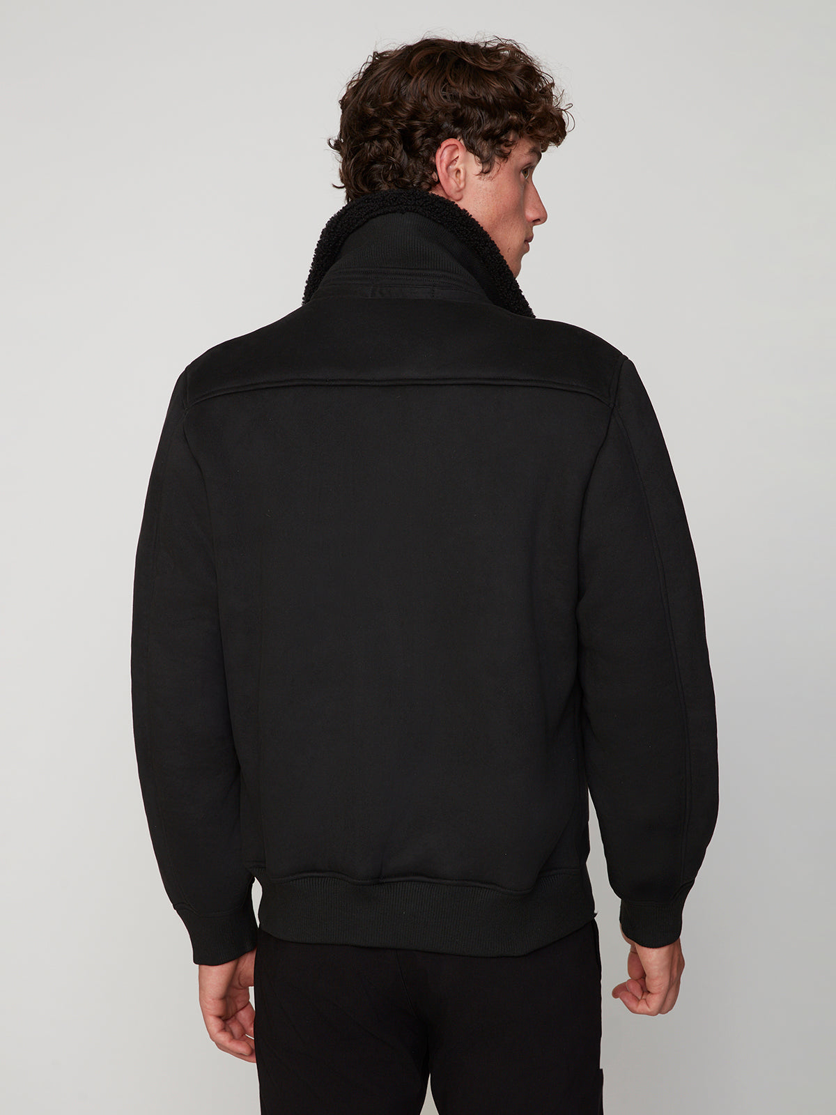 A man with curly brown hair, seen from the back, wears a black PROJEK RAW MENS BONDED MICRO SUEDE JACKET with a high collar and black pants against a plain light gray background.