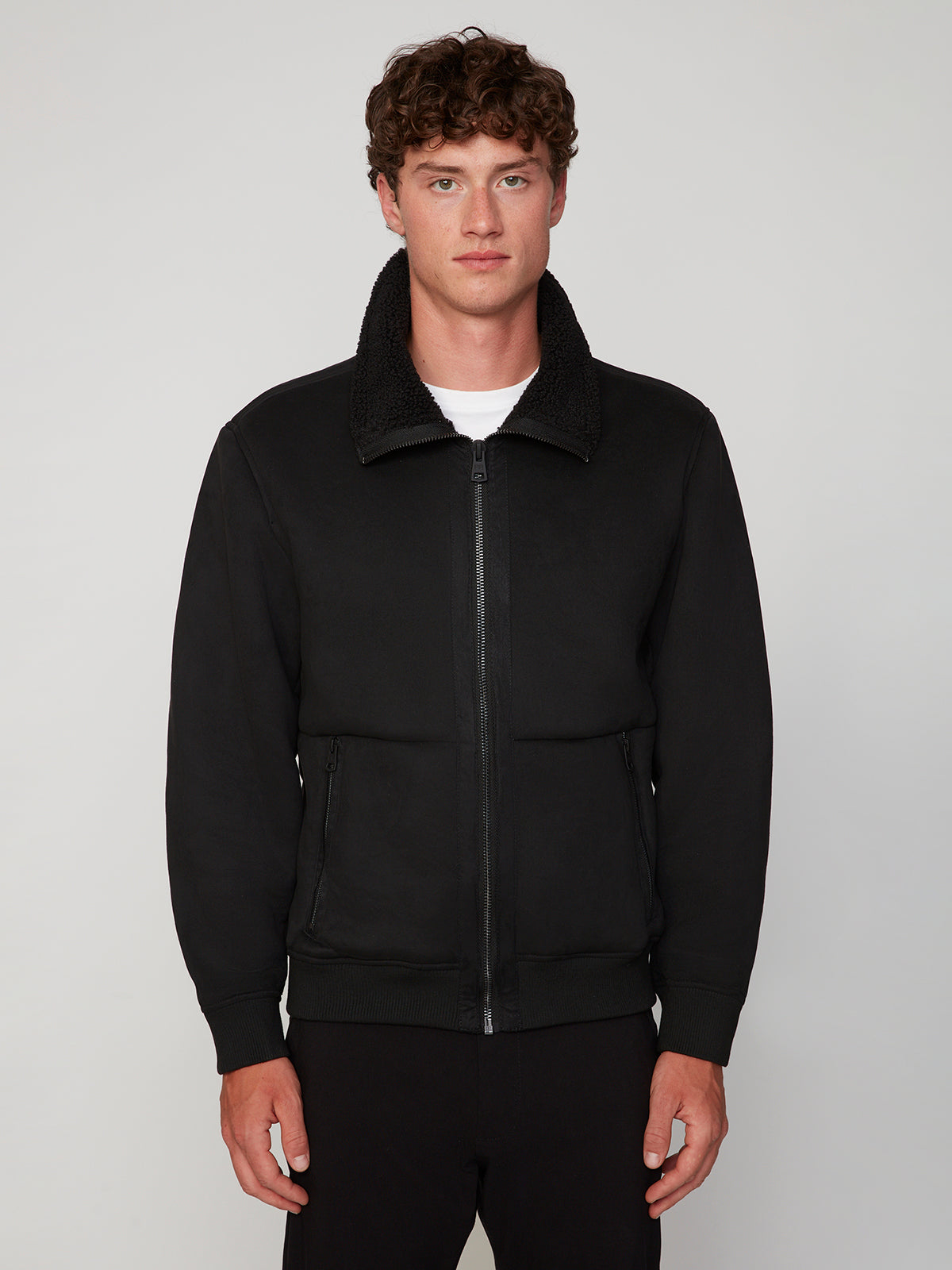 A young man with curly brown hair faces forward, wearing the PROJEK RAW MENS BONDED MICRO SUEDE JACKET in black and black pants, posed against a plain light gray background.