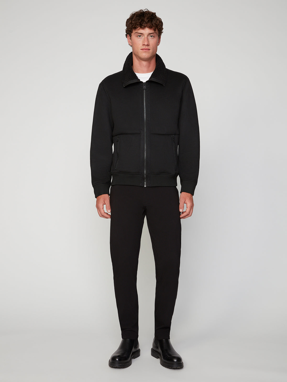 A man stands facing forward against a plain light background, wearing the PROJEK RAW MENS BONDED MICRO SUEDE JACKET in black, paired with black pants, black boots, and a white t-shirt underneath.