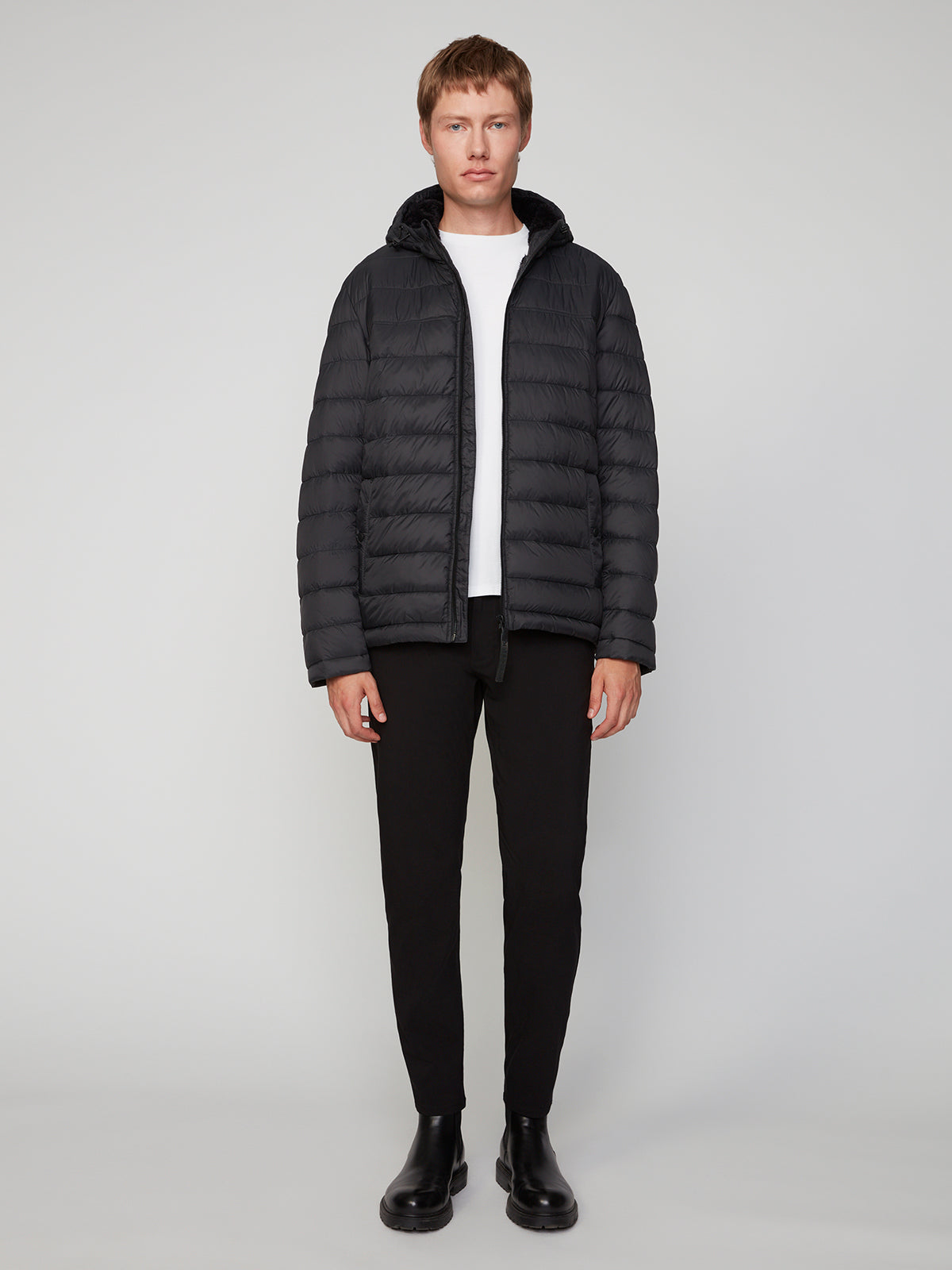 A person stands facing forward, wearing the PROJEK RAW MENS HOODED FAUX FUR LINED PUFFER JACKET over a white shirt, black pants, and black boots against a plain light gray background—a stylish cold-weather essential.