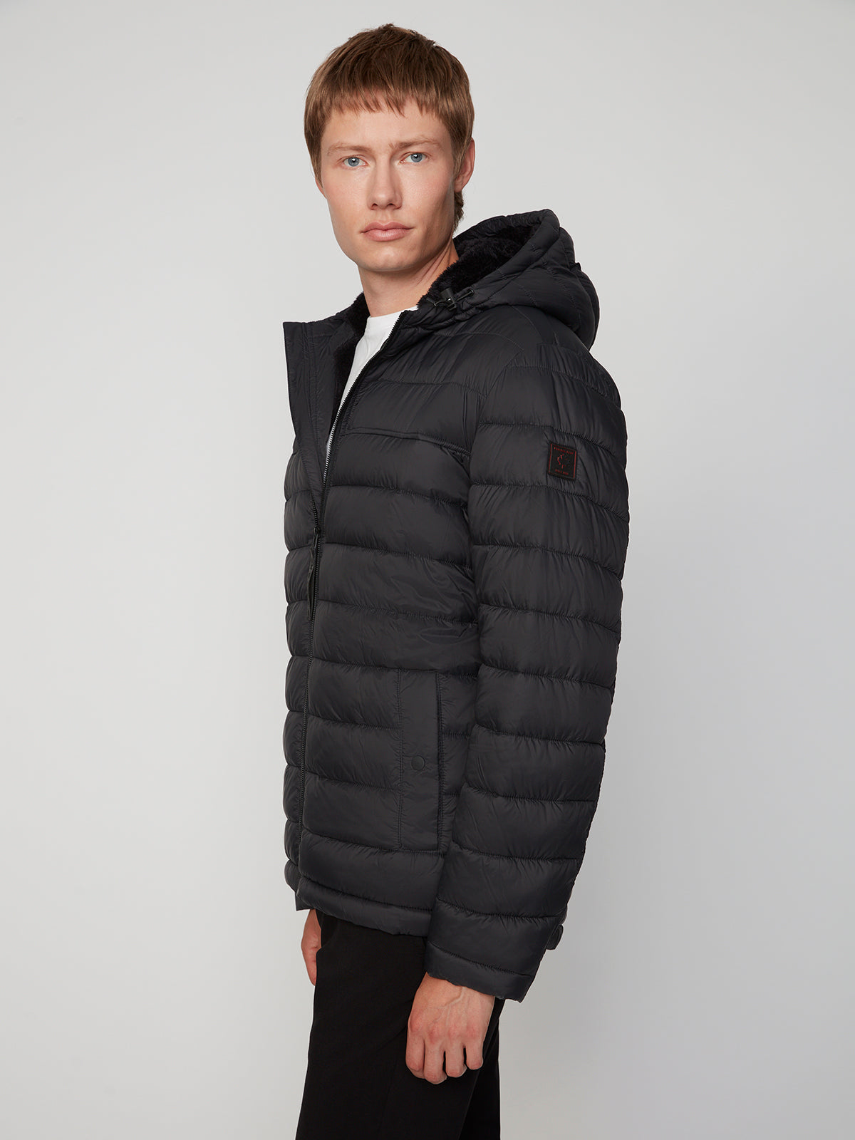 A man with short light brown hair wears the PROJEK RAW MENS HOODED FAUX FUR LINED PUFFER JACKET and black pants, standing slightly turned to the side against a plain light gray background.