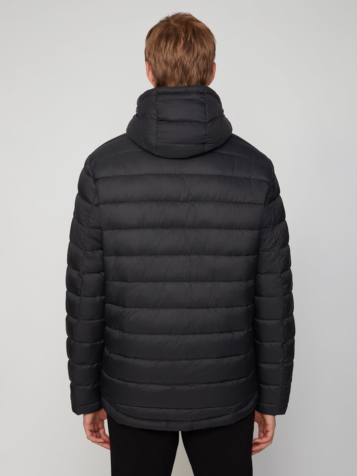 A person with light brown hair is shown from behind, wearing the PROJEK RAW MENS HOODED FAUX FUR LINED PUFFER JACKET with horizontal quilting, standing against a plain light grey background—the ideal cold-weather choice.