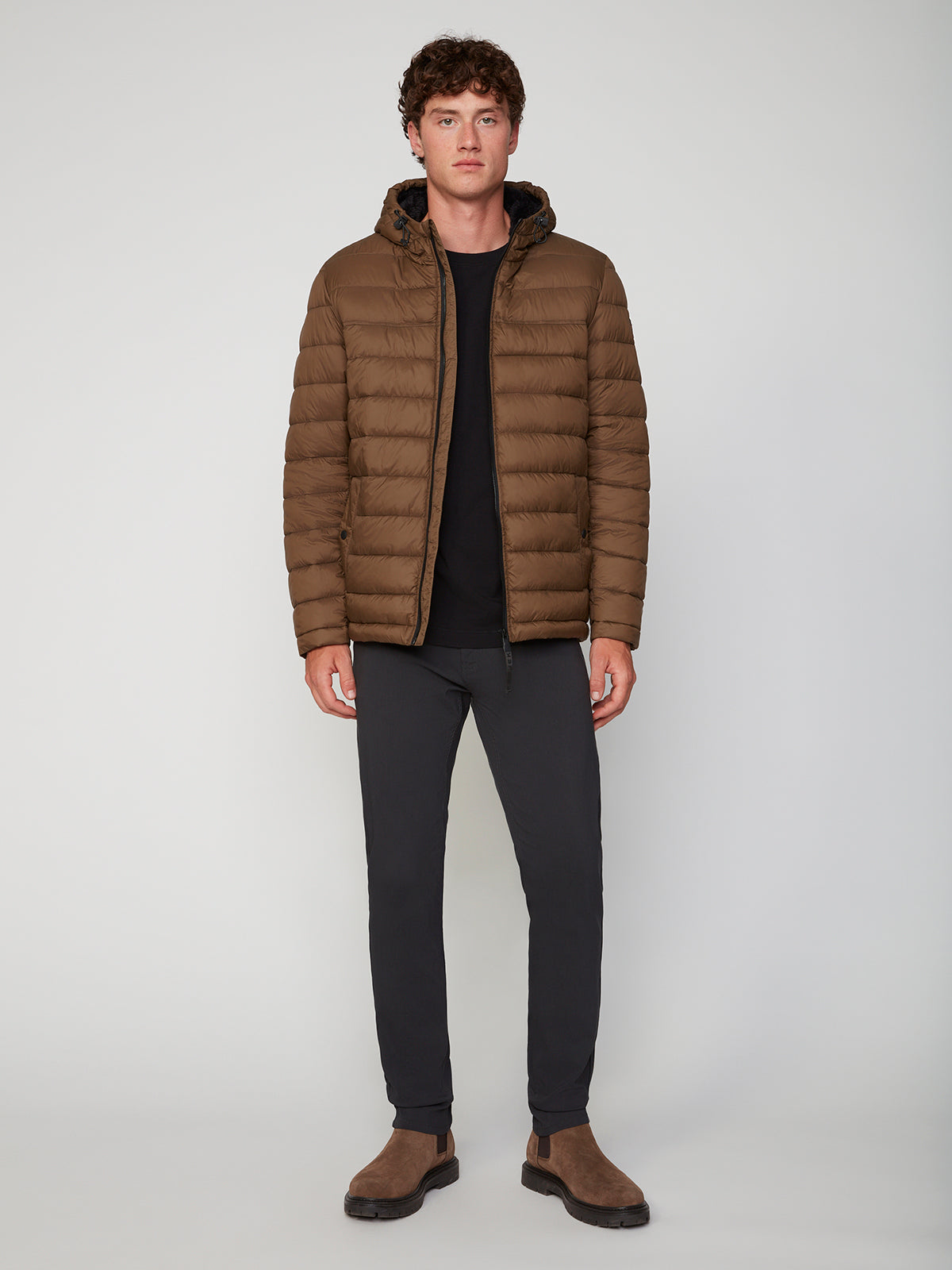 A person stands against a plain background wearing the PROJEK RAW MENS HOODED FAUX FUR LINED PUFFER JACKET for warmth, paired with a black shirt, dark pants, and brown suede boots. The jacket is zipped and hooded; they face forward with a neutral expression.