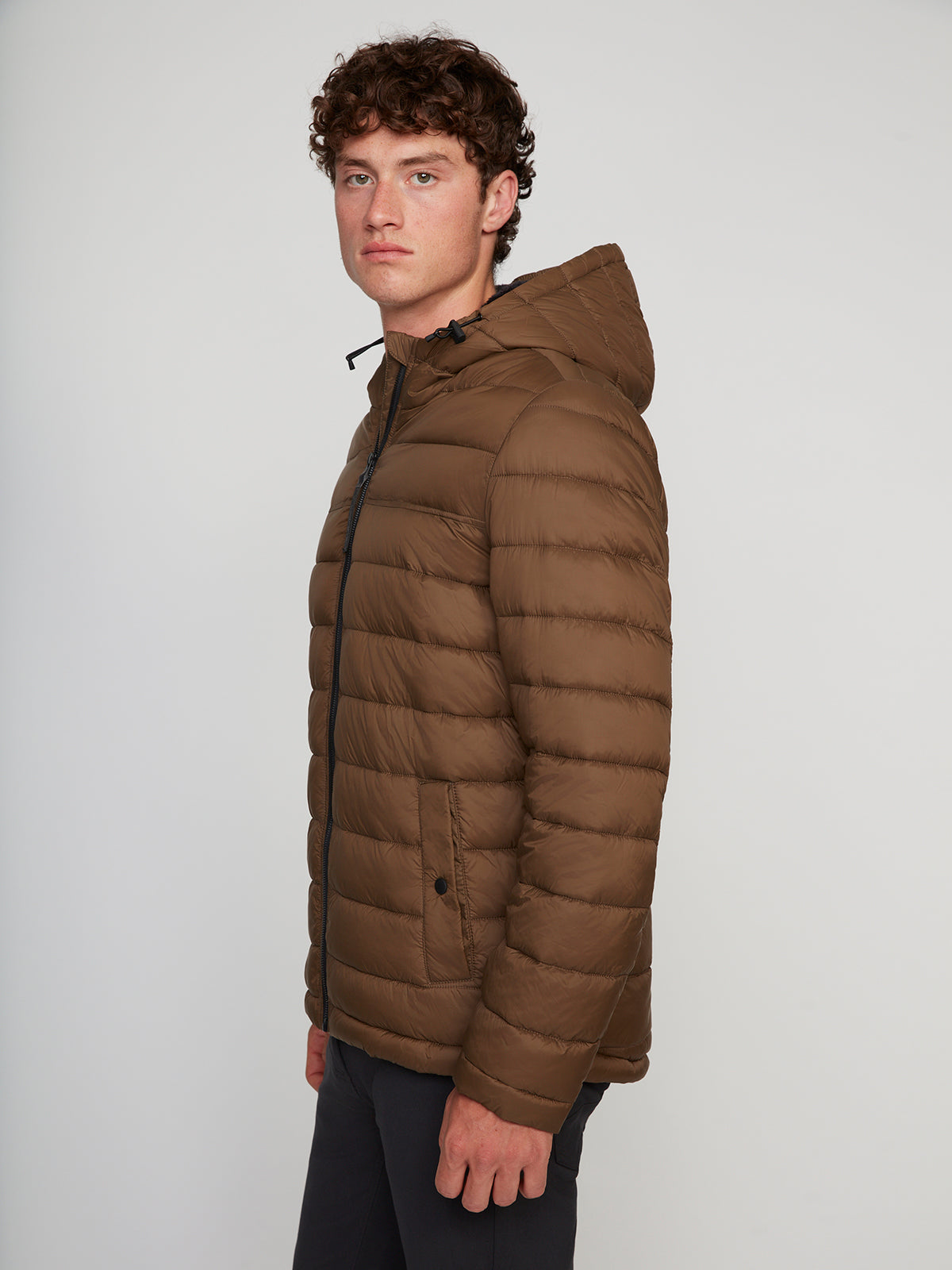 A young man with curly hair stands sideways, showcasing the cold-weather comfort of the PROJEK RAW MENS HOODED FAUX FUR LINED PUFFER JACKET with horizontal quilting and black pants, against a plain light background.