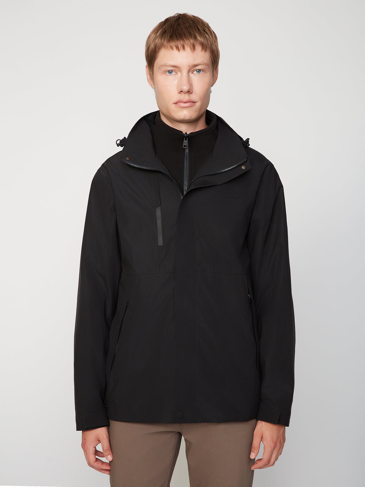 A person with short light brown hair wears the PROJEK RAW MENS 3 IN 1 WATERPROOF JACKET in black over a black zip-up top and brown pants, standing against a plain light gray background.