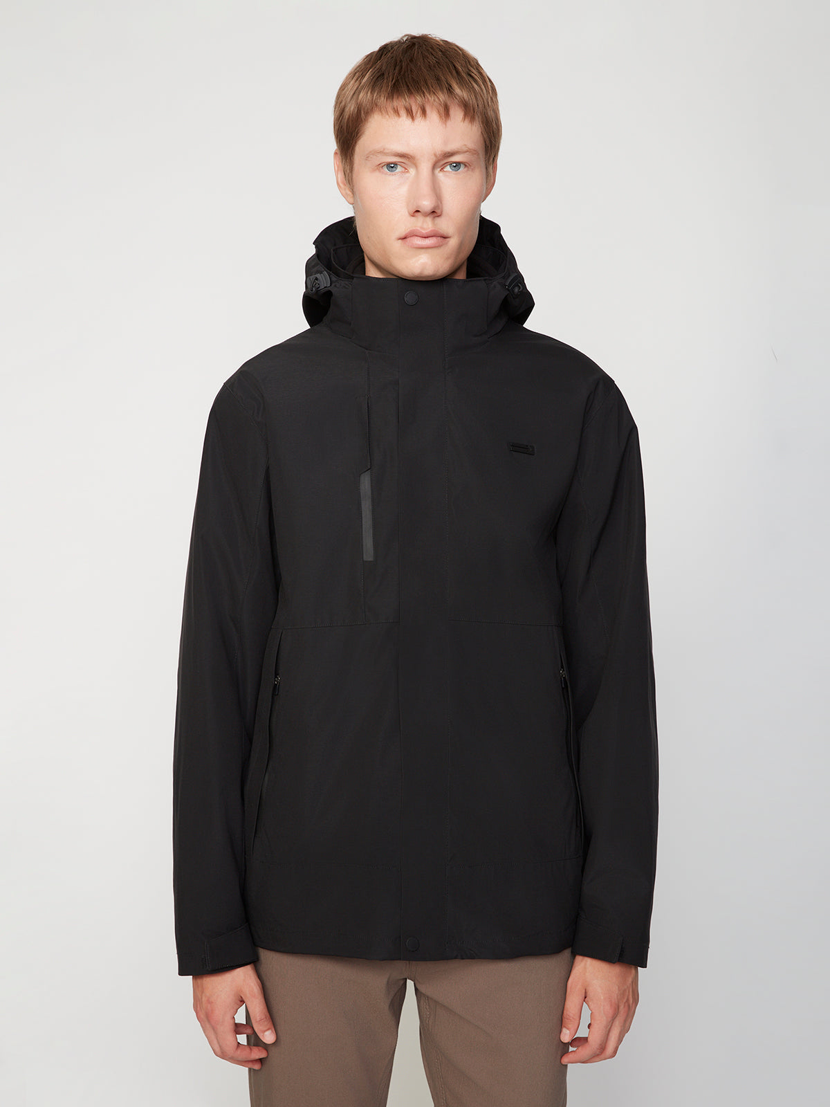 A person with short light brown hair wears a PROJEK RAW MENS 3 IN 1 WATERPROOF JACKET with a hood and brown pants, standing facing forward against a plain, light background.