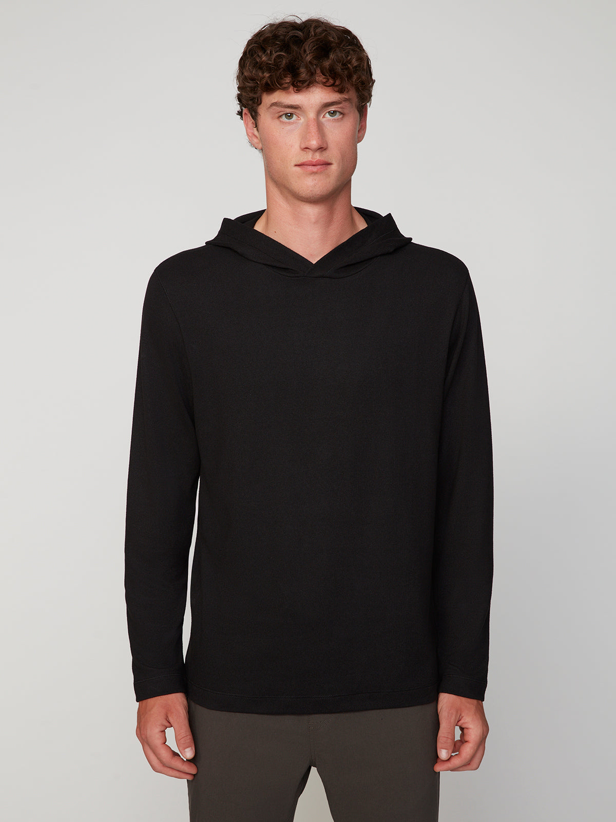 A young man with curly brown hair stands against a light background, wearing the PROJEK RAW MENS HOODED LONG SLEEVE KNIT and dark pants, looking straight at the camera with a neutral expression—perfect for layering.