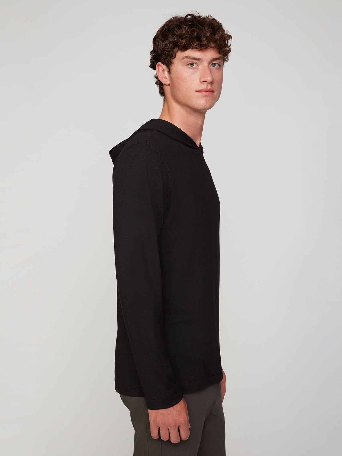 A young man with curly brown hair wears the PROJEK RAW MENS HOODED LONG SLEEVE KNIT and gray pants, standing sideways and looking at the camera against a plain light background—perfect for stylish layering.