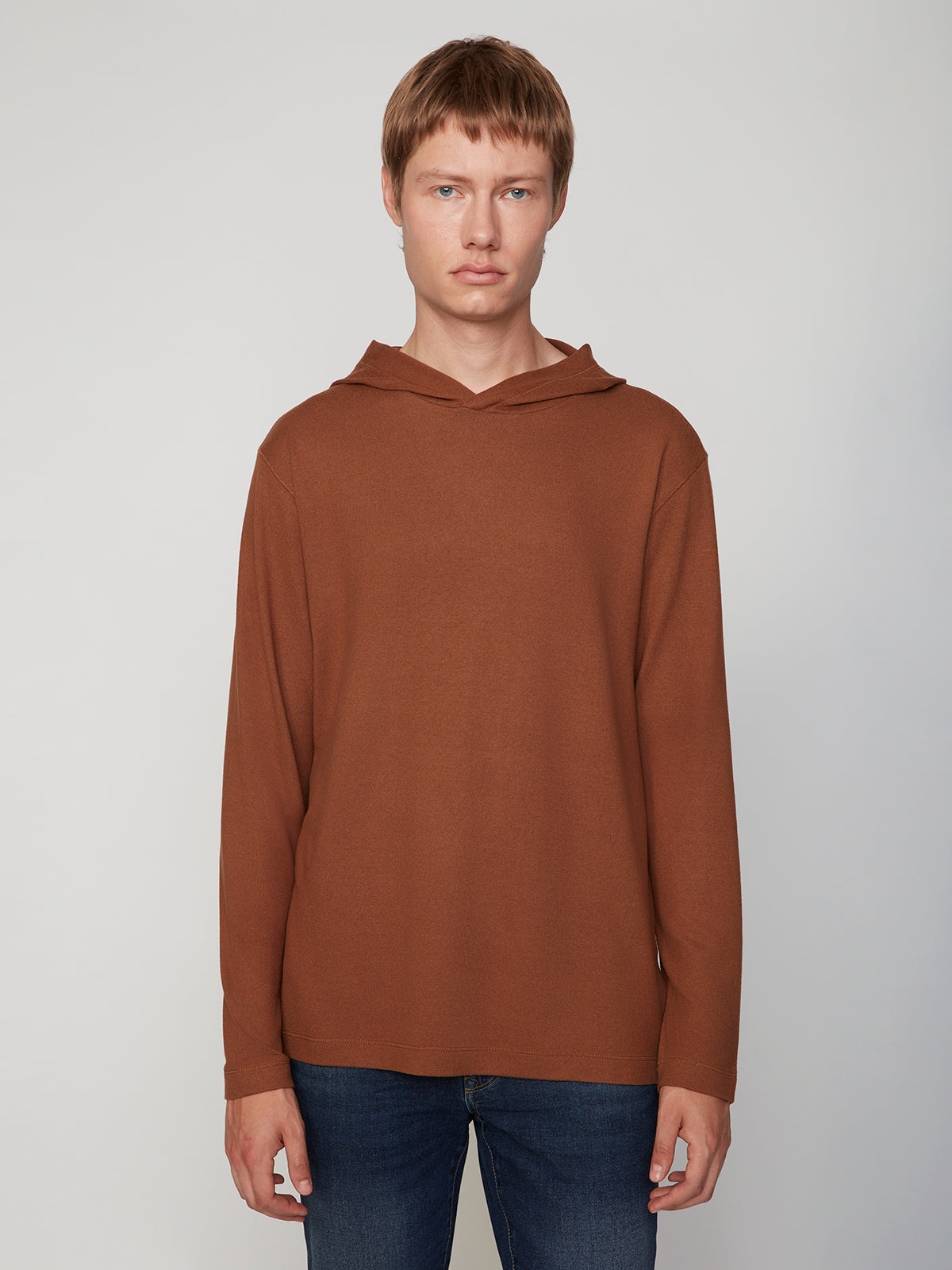 A person with short light brown hair wears the PROJEK RAW MENS HOODED LONG SLEEVE KNIT and blue jeans, standing against a plain light gray background, facing forward with a neutral expression for casual comfort.