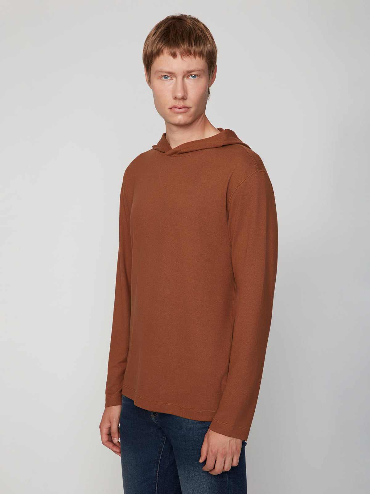 A young man with short light brown hair models the PROJEK RAW MENS HOODED LONG SLEEVE KNIT and blue jeans, standing against a plain light gray background and looking directly at the camera with a neutral expression.